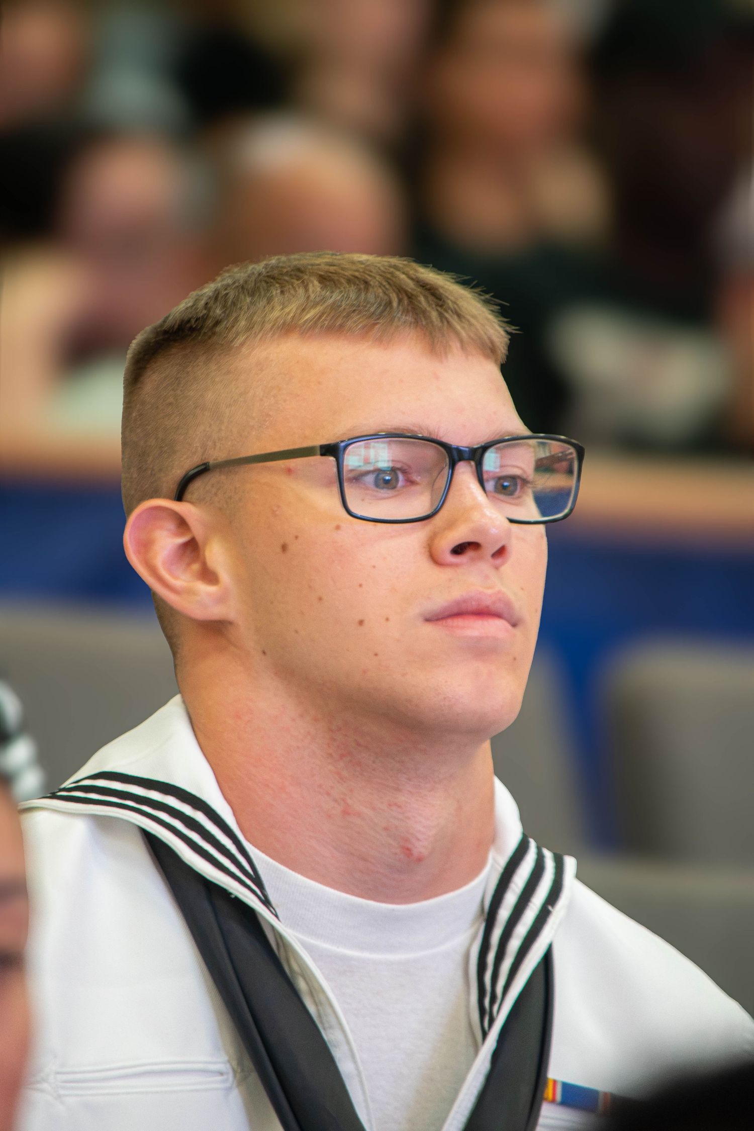 Young Navy sailor in dress whites with glasses attends a formal military ceremony.