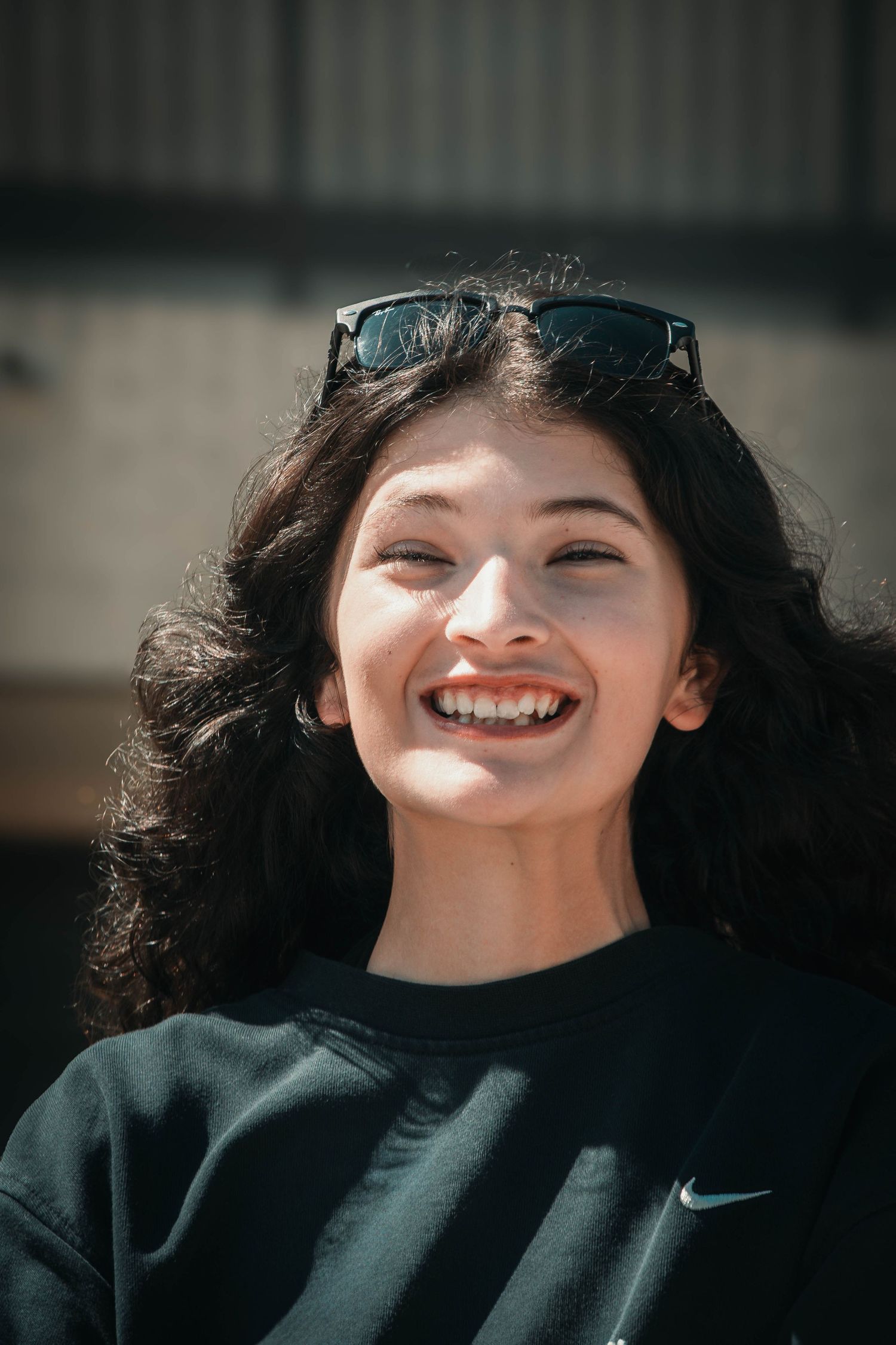 Young woman with curly black hair and sunglasses on head, smiling brightly outdoors in a black Nike sweatshirt.