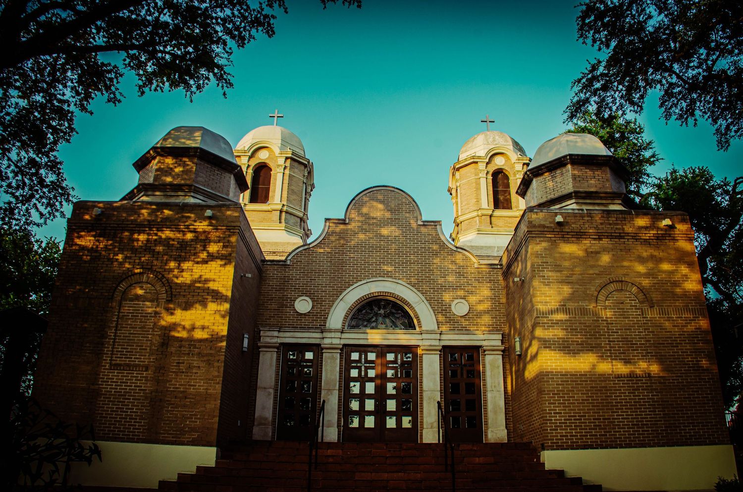 Historic brick church with domed towers and ornate facade framed by trees under a blue sky.