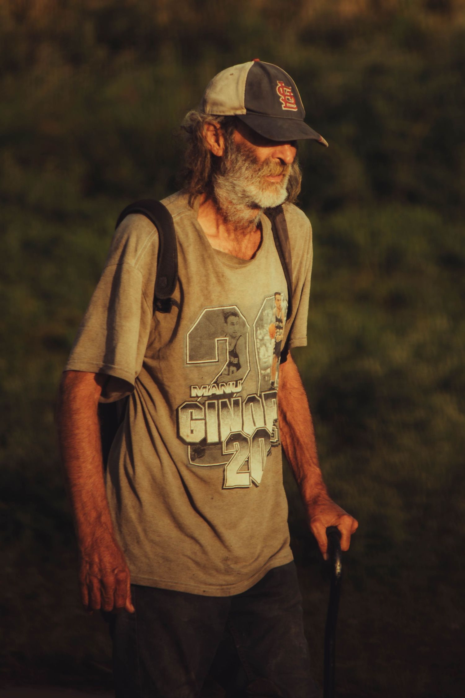 An elderly homeless man with a beard wearing a baseball cap and backpack walking outdoors at sunset.