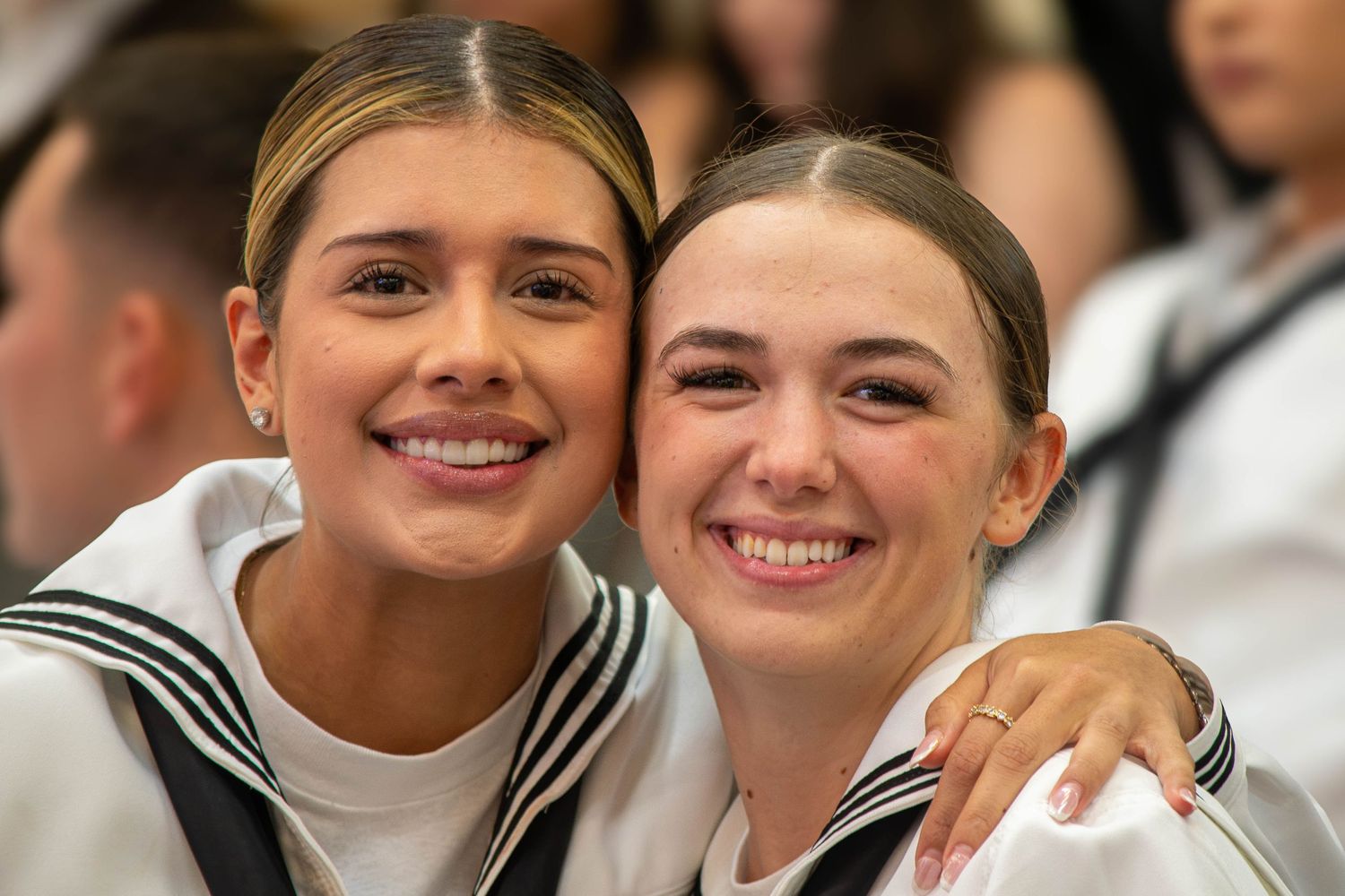 Two smiling female Navy sailors in white uniforms pose together at a military graduation ceremony.