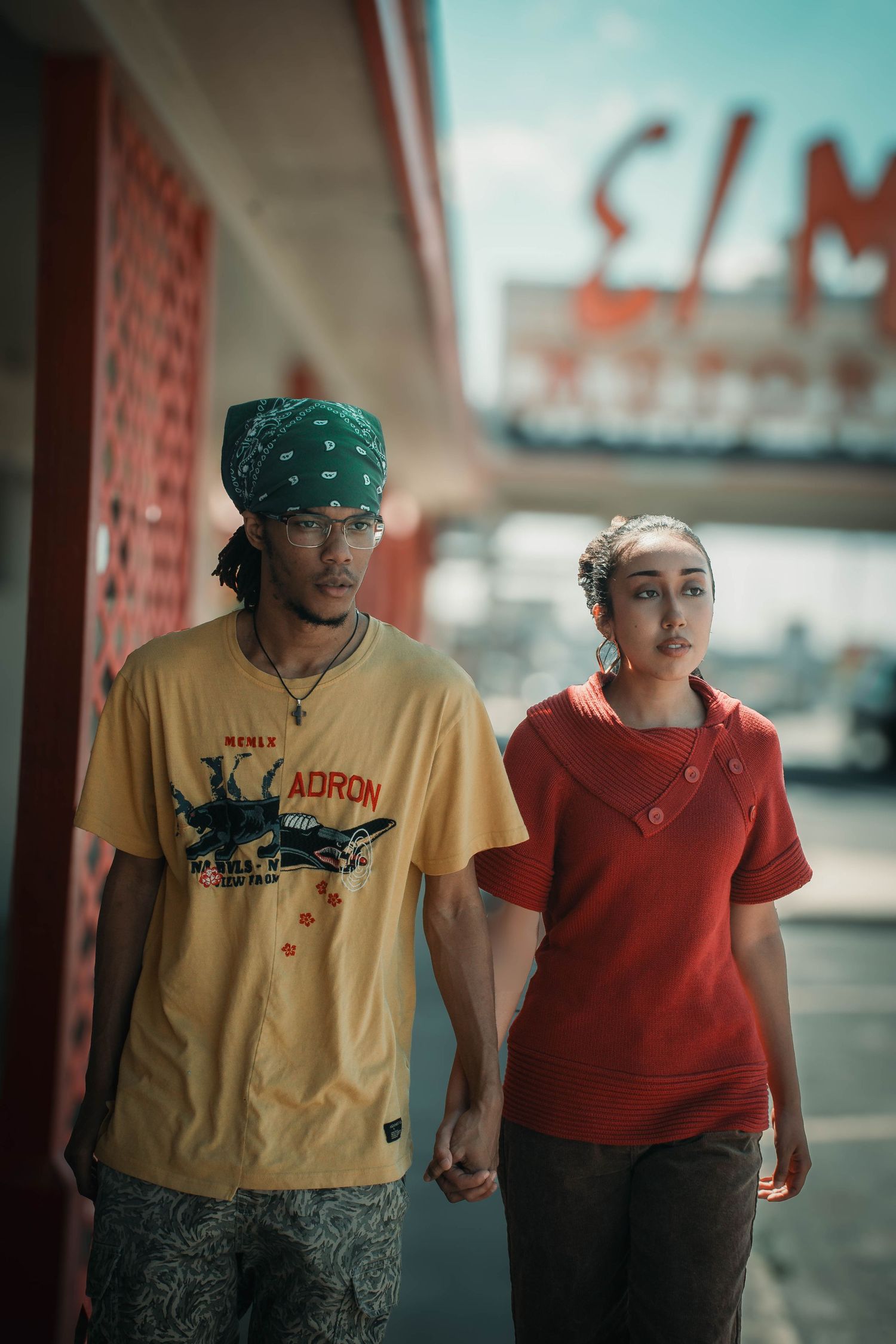 A young couple holding hands outside a retro cinema, man in yellow ADRON tee and green bandana, woman in red top.