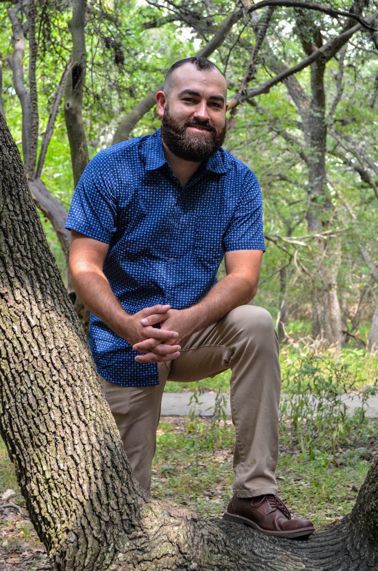 Bearded man in blue shirt sitting on tree roots in a forest setting.