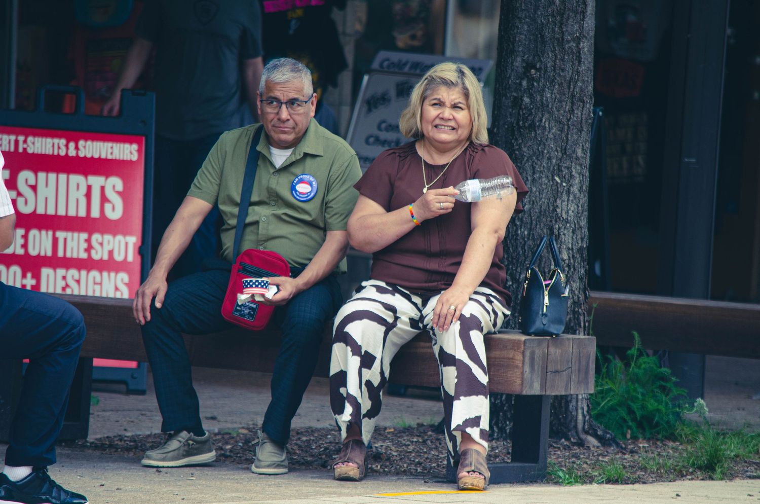 Two people sitting on a bench outside a t-shirt souvenir shop in a casual outdoor setting.