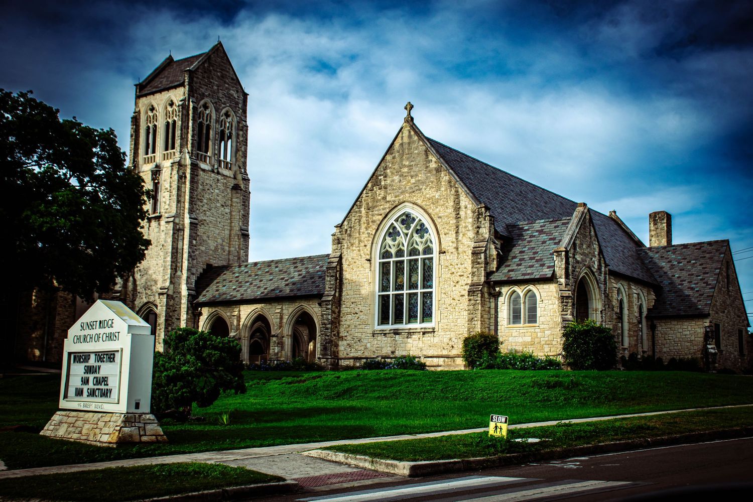 Historic stone Gothic Revival church with bell tower and arched windows under dramatic cloudy sky.