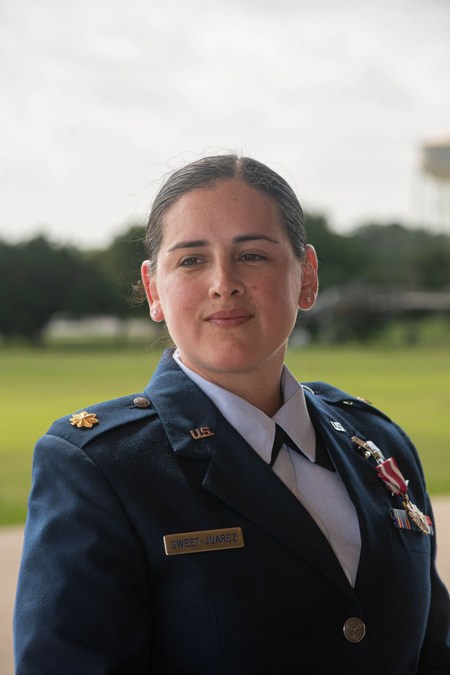 A female US Air Force officer in dress uniform with medals smiles outdoors on a airfield.