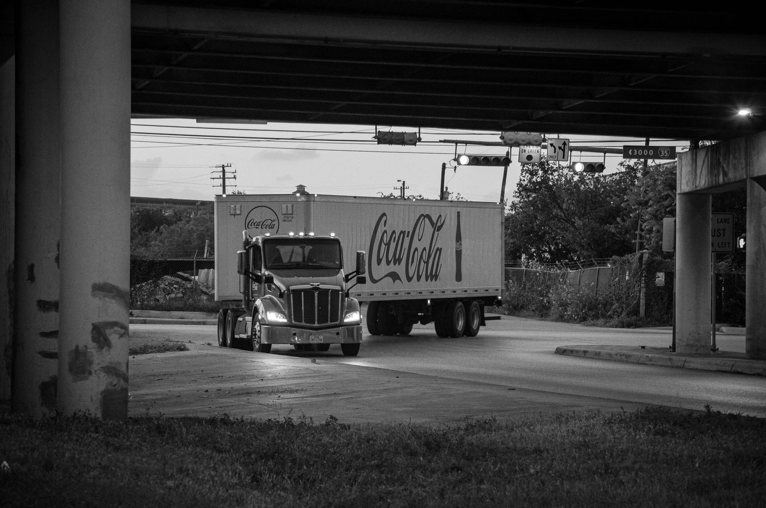 Black and white photo of a Coca-Cola delivery truck driving under an overpass at dusk.