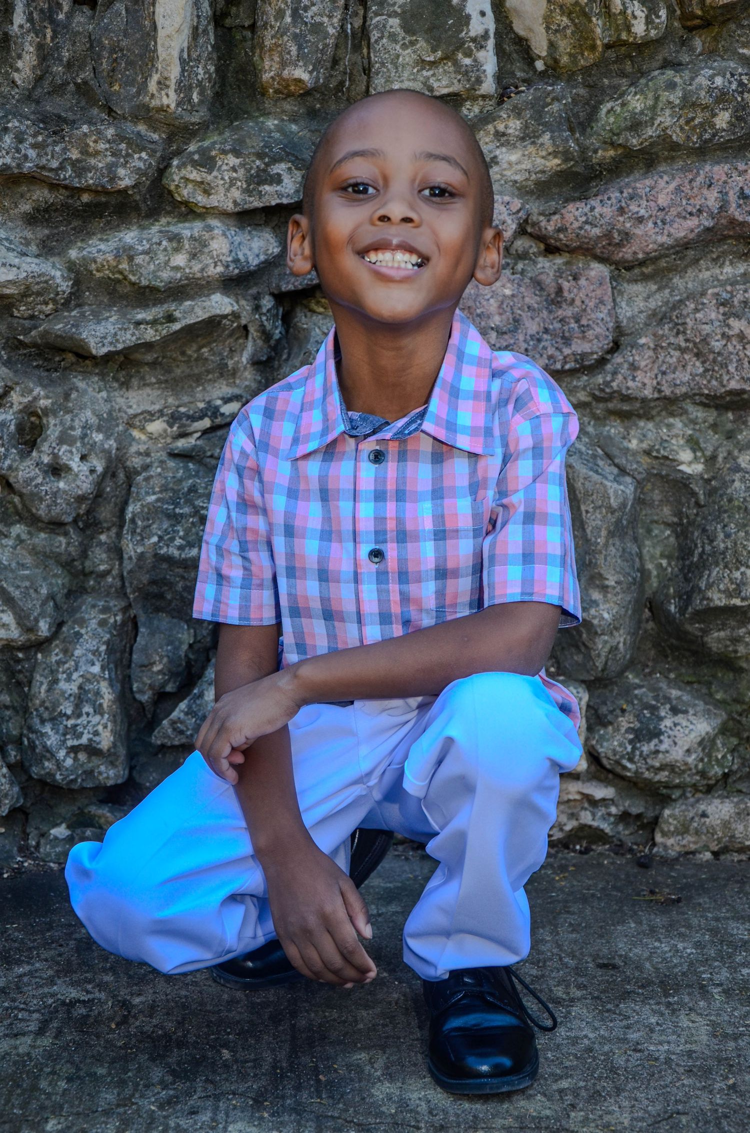 Smiling young Black boy in plaid shirt crouching against a stone wall.