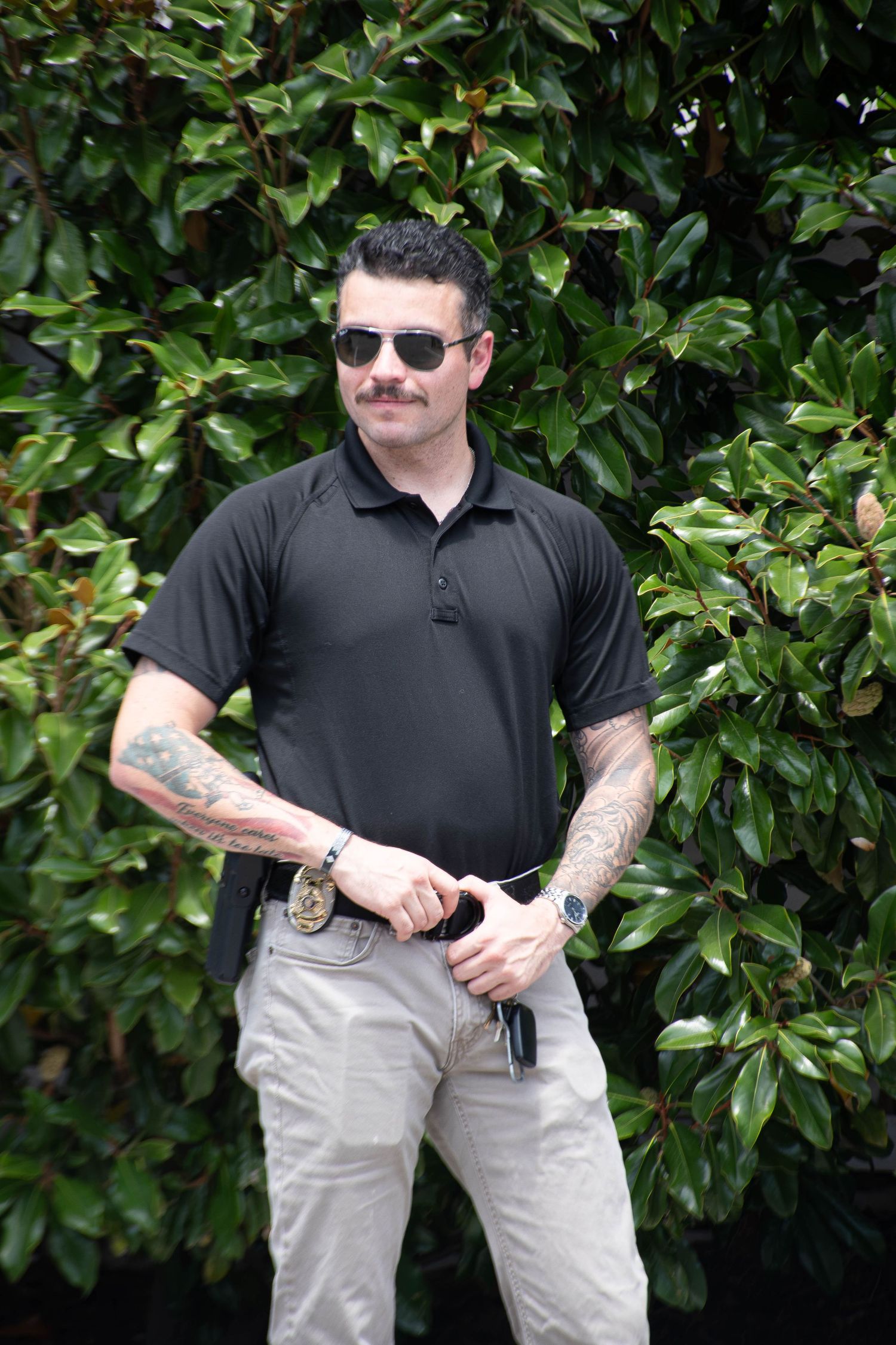 Tattooed man with mustache in black polo shirt and sunglasses standing before green foliage outdoors.