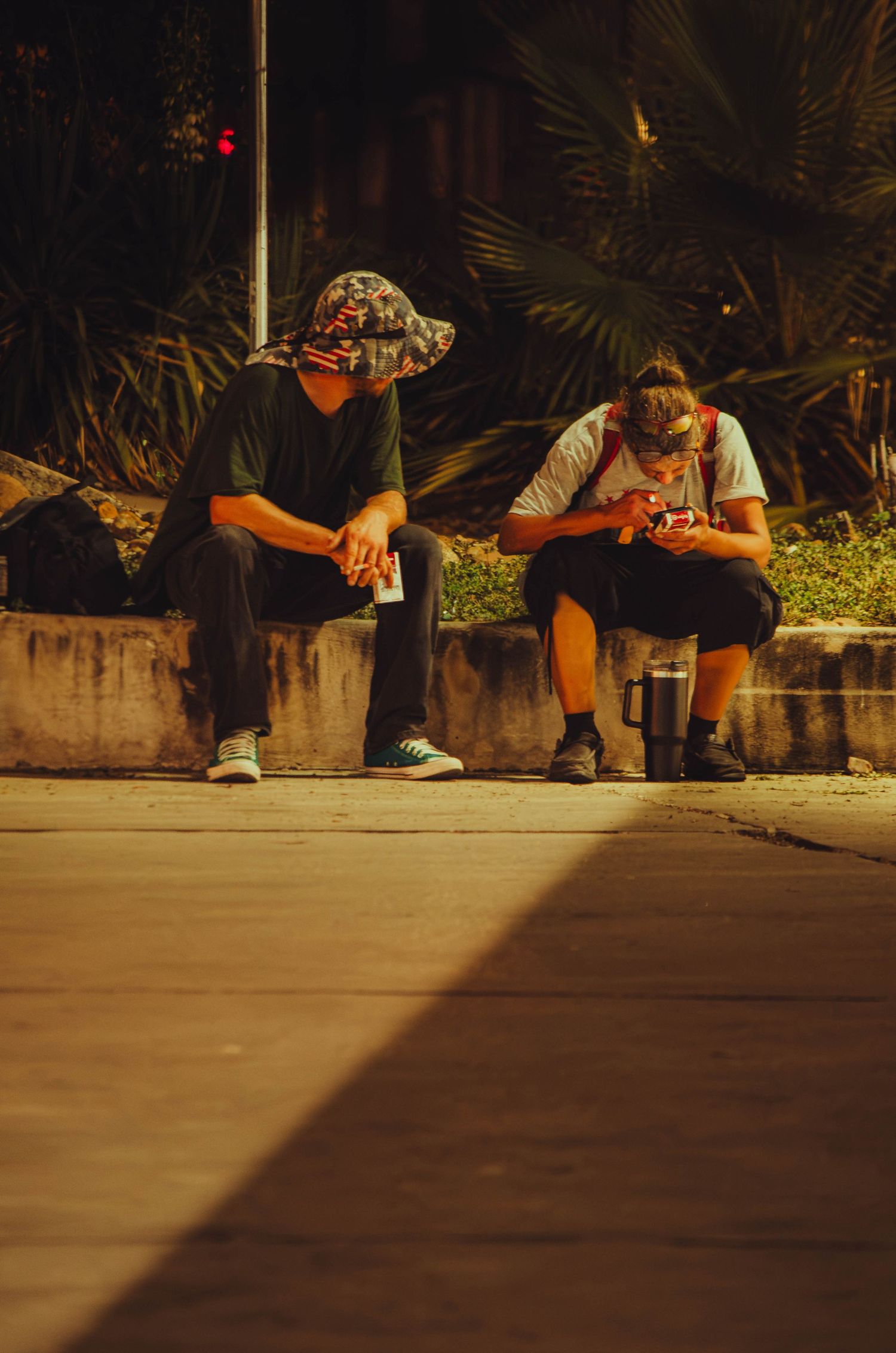 Two young men wearing masks and hats sitting on a curb at night looking at their phones.