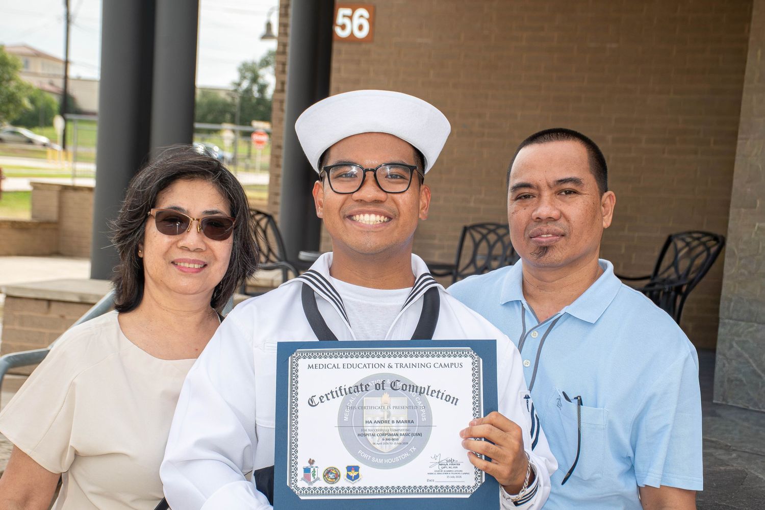 Navy sailor in white uniform proudly holds Certificate of Completion flanked by proud parents after military graduation.