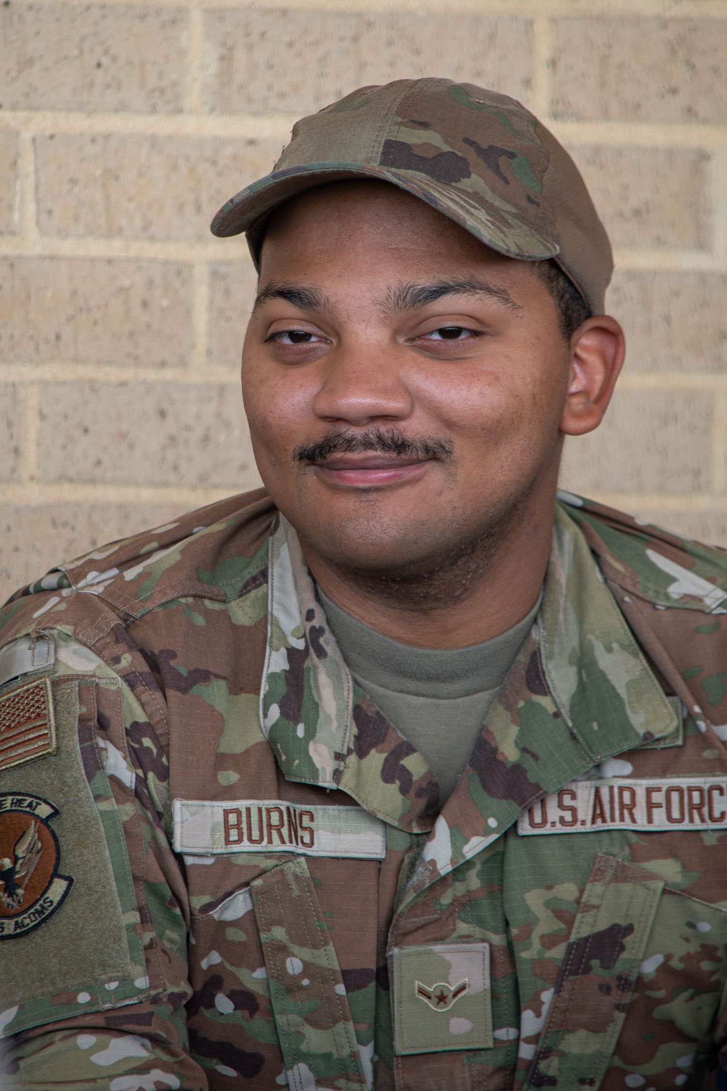 US Air Force airman Burns in camouflage uniform and cap smiles against a brick wall.