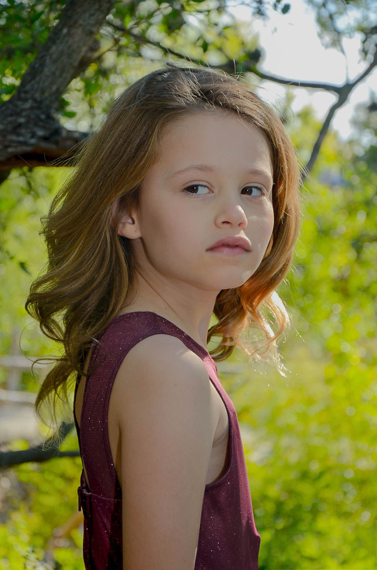 Young girl with flowing auburn hair in a purple sleeveless dress gazes back under a sunlit tree.