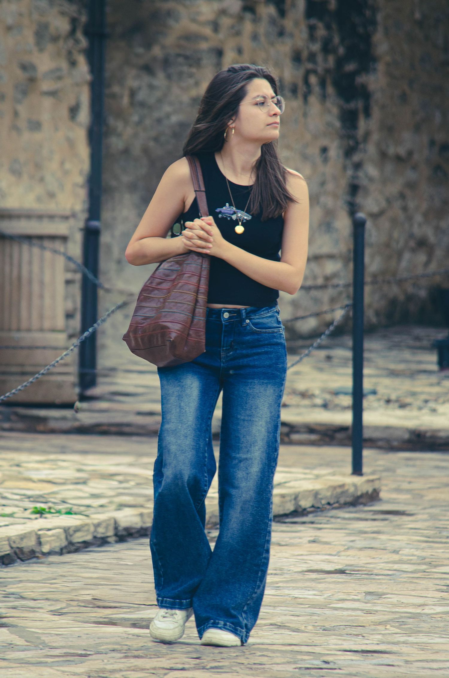 Young woman in wide-leg jeans and black tank top carrying a brown leather bag on a cobblestone street.