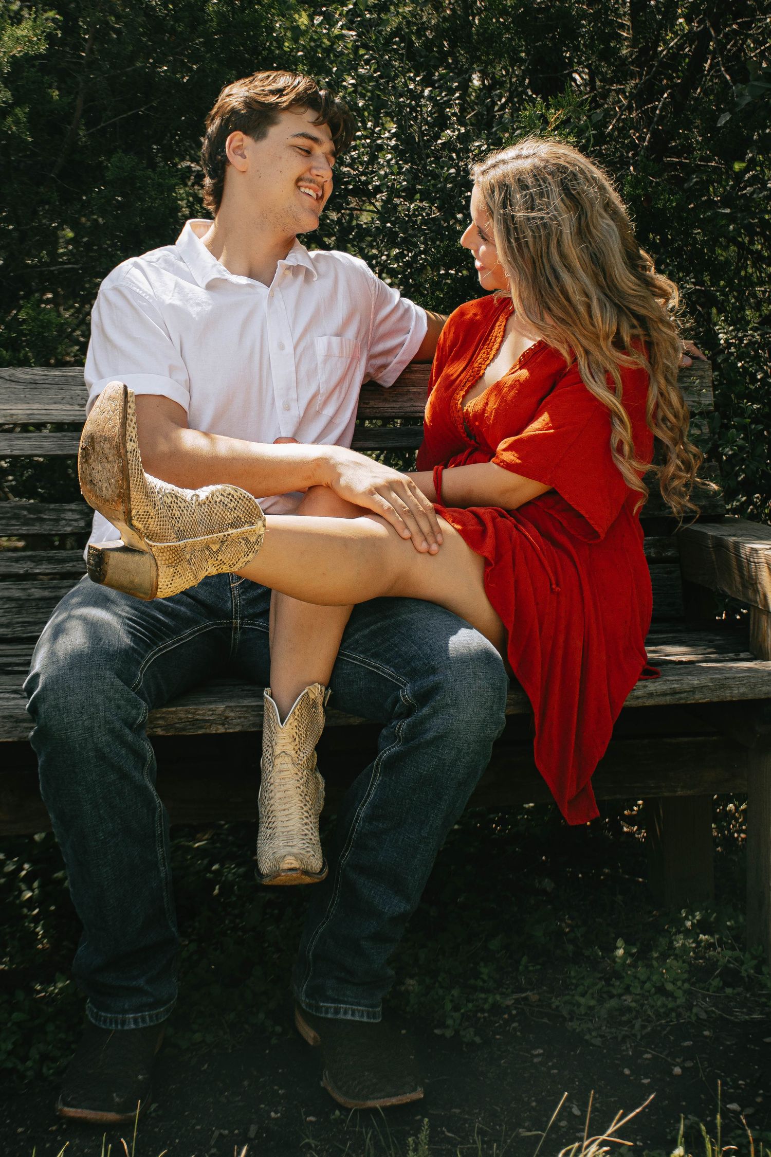 Couple sitting on a wooden bench laughing together, woman wearing cowboy boots and red dress.