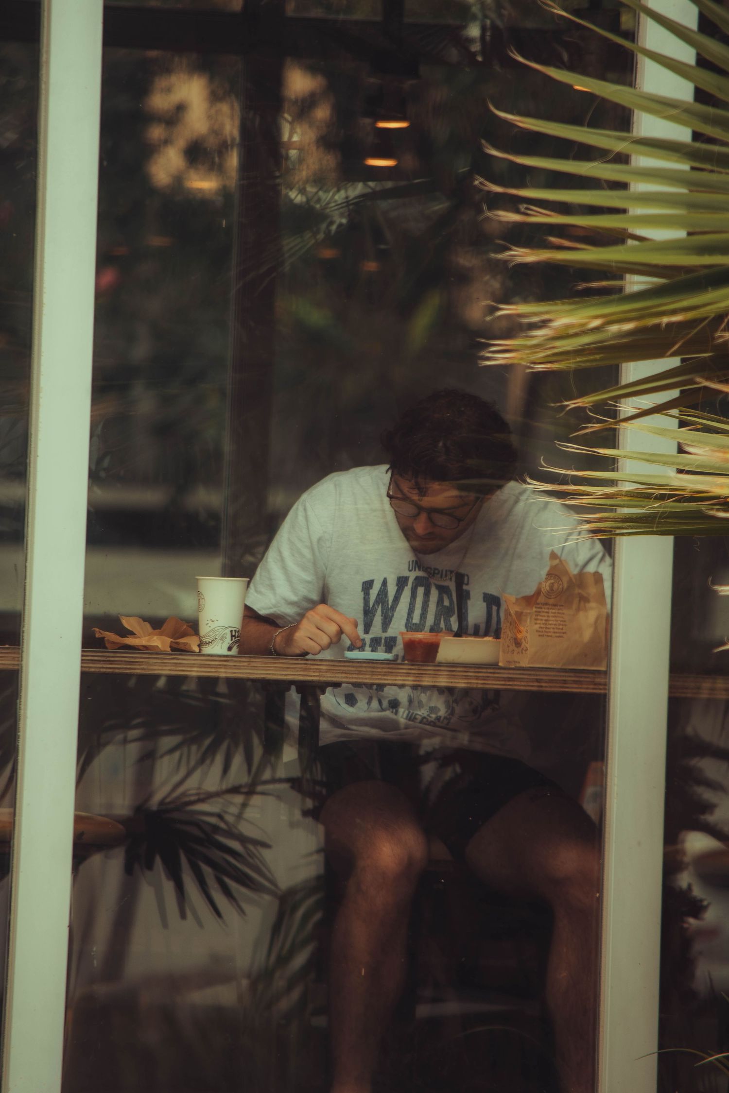 A young man in a World t-shirt studying at a cafe table, seen through a glass window.