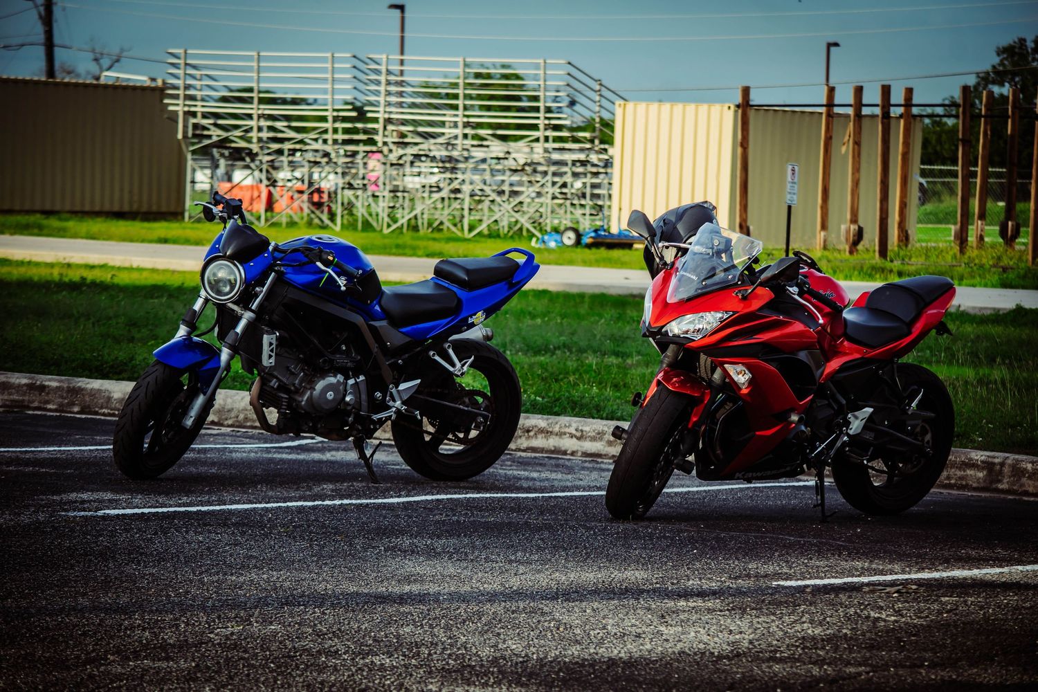 Two motorcycles, a blue naked bike and a red sport bike, parked side by side in a lot near a grassy field.