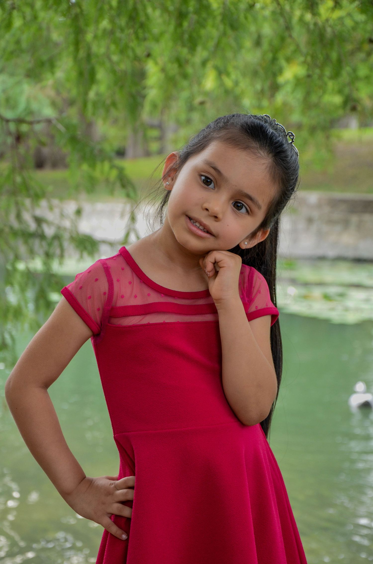 Young girl in red dress posing near pond with willow tree in background at park.