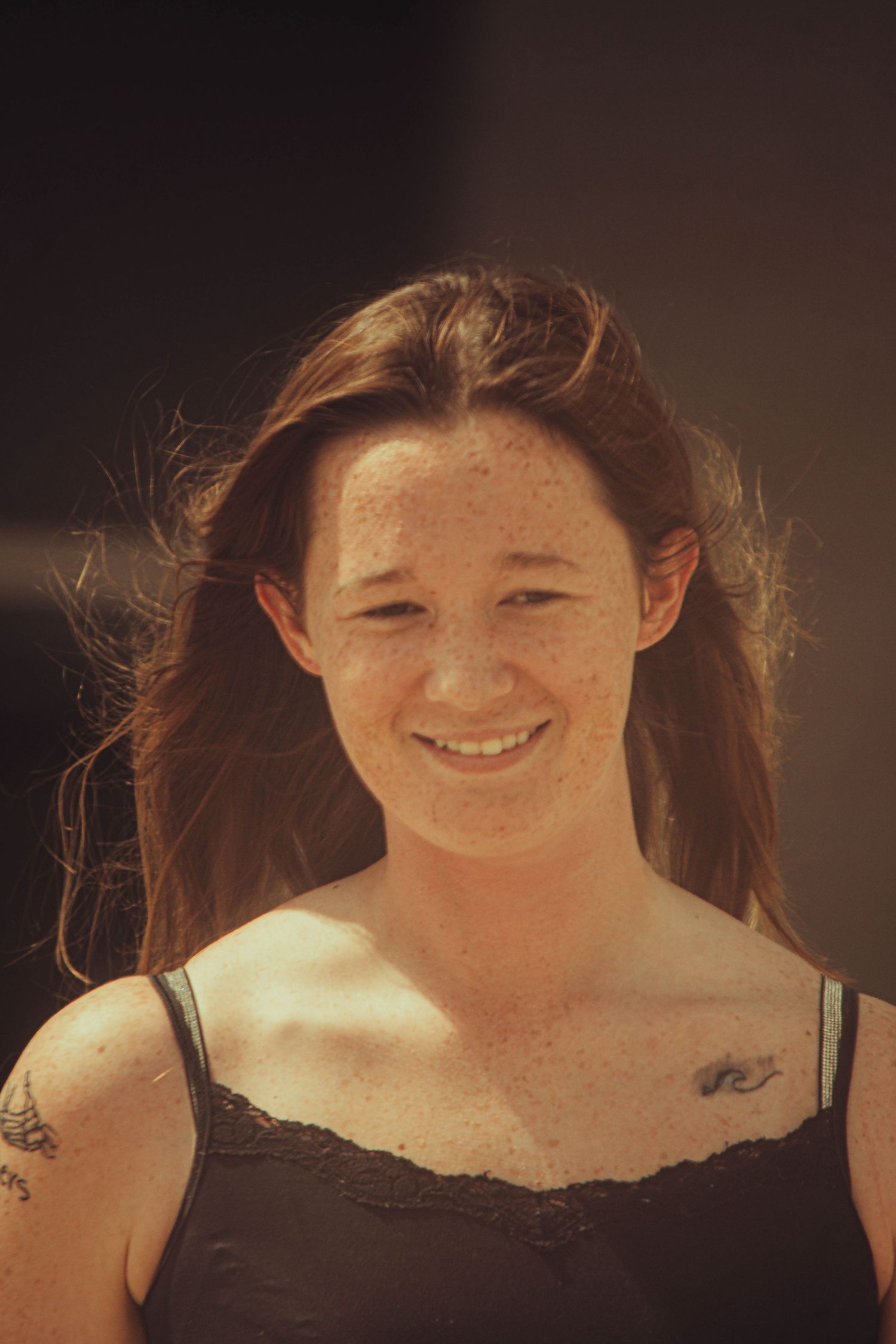 Smiling young woman with freckles and auburn hair wearing a black tank top with tattoos outdoors in sunlight.
