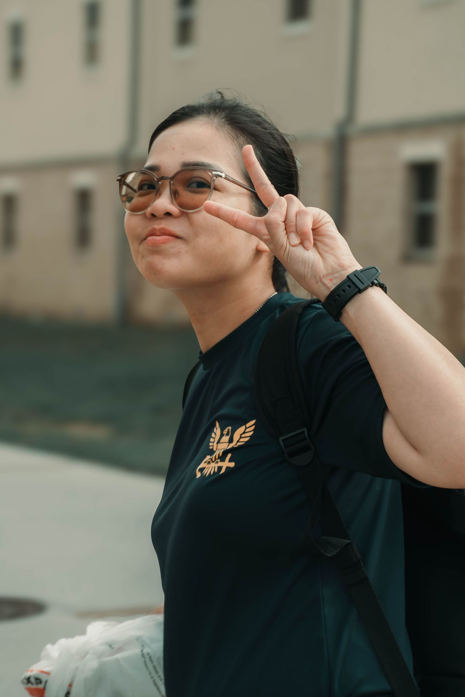 Asian woman in black tee flashing peace sign wearing glasses and backpack outdoors.