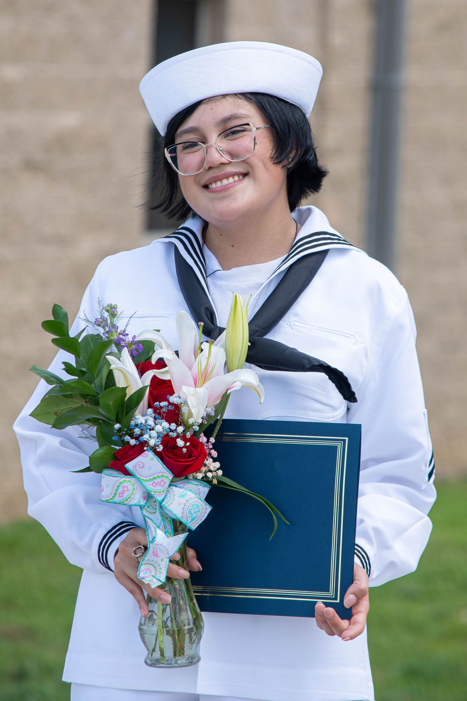 Smiling US Navy sailor in white dress uniform holding flowers and a diploma after graduation.