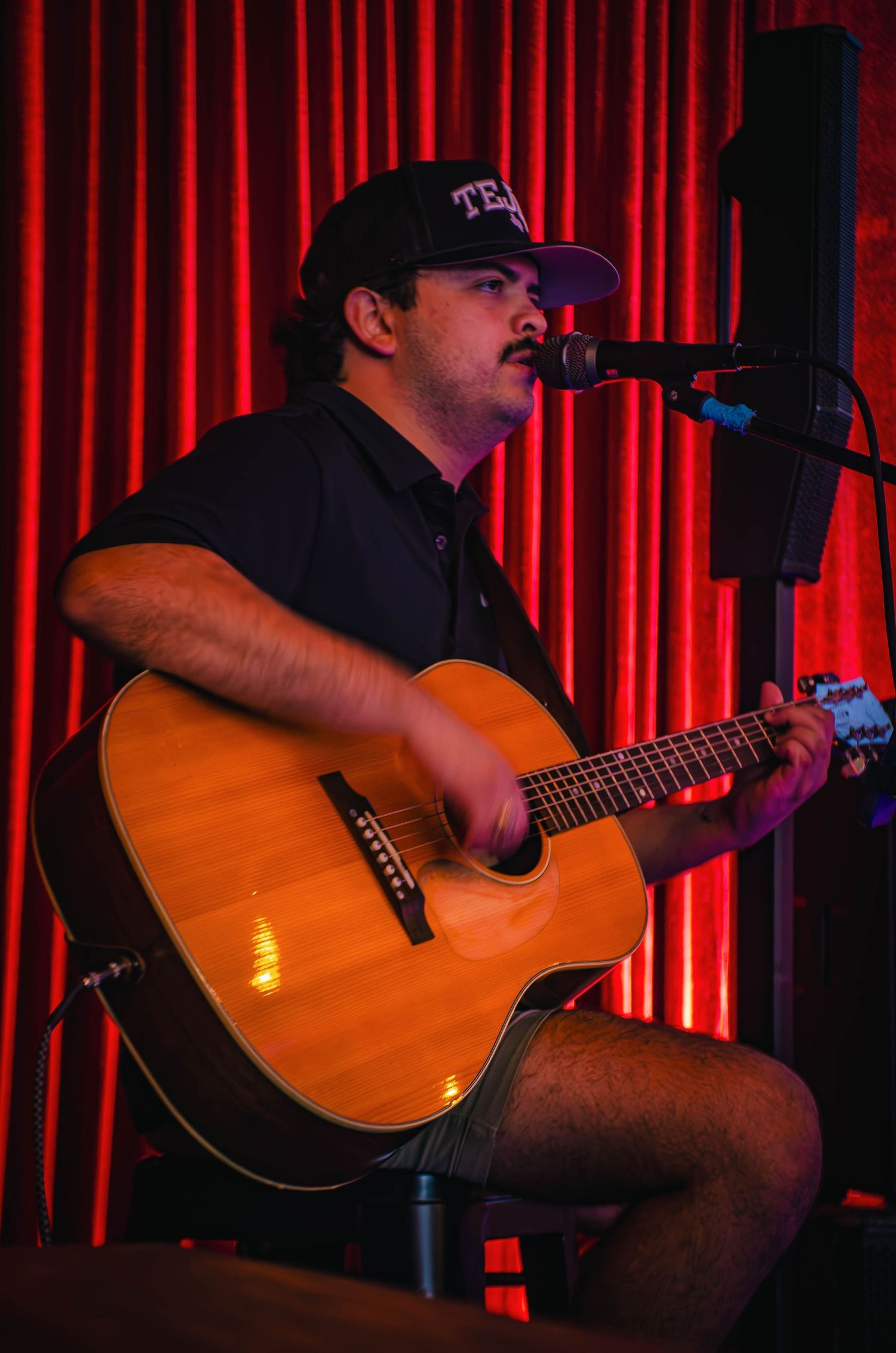 Male musician performing on acoustic guitar and singing into microphone on a red-lit stage.