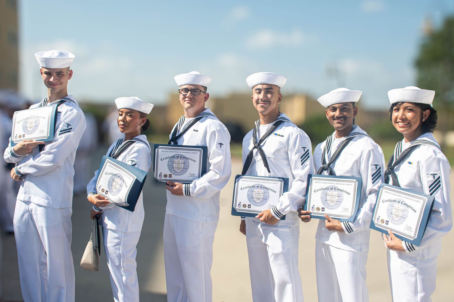 Six Navy sailors in white dress uniforms proudly display certificates of achievement at outdoor military graduation.