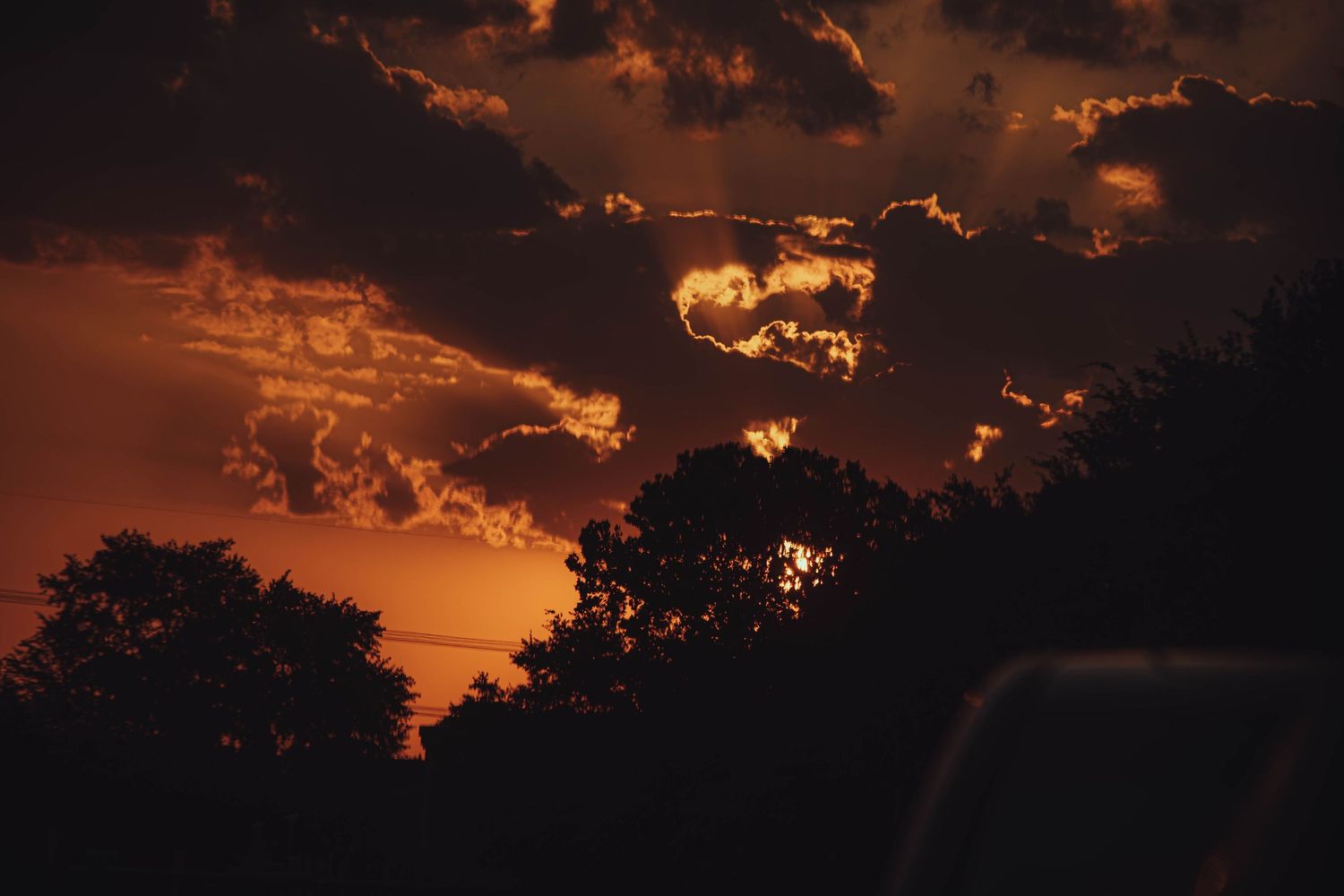 Fiery orange and red sunset sky with dark tree silhouettes and glowing cloud formations.