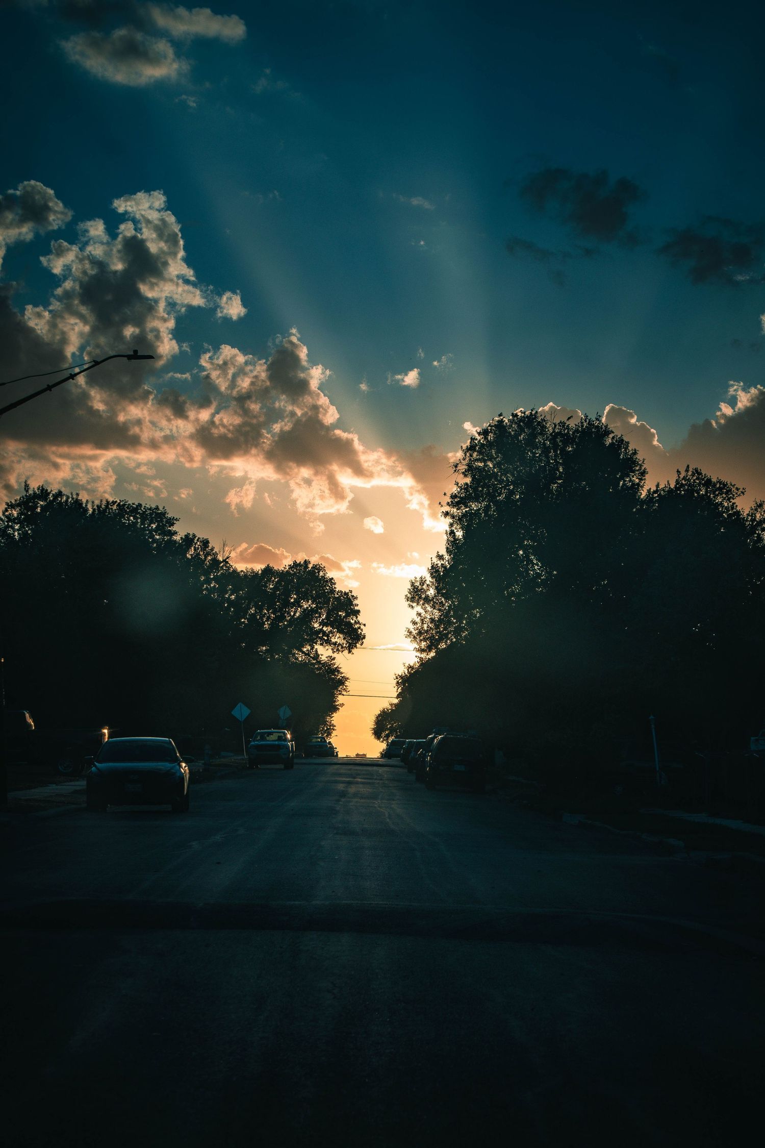 Dramatic sunset with sun rays breaking through clouds over a tree-lined suburban street with silhouetted cars.