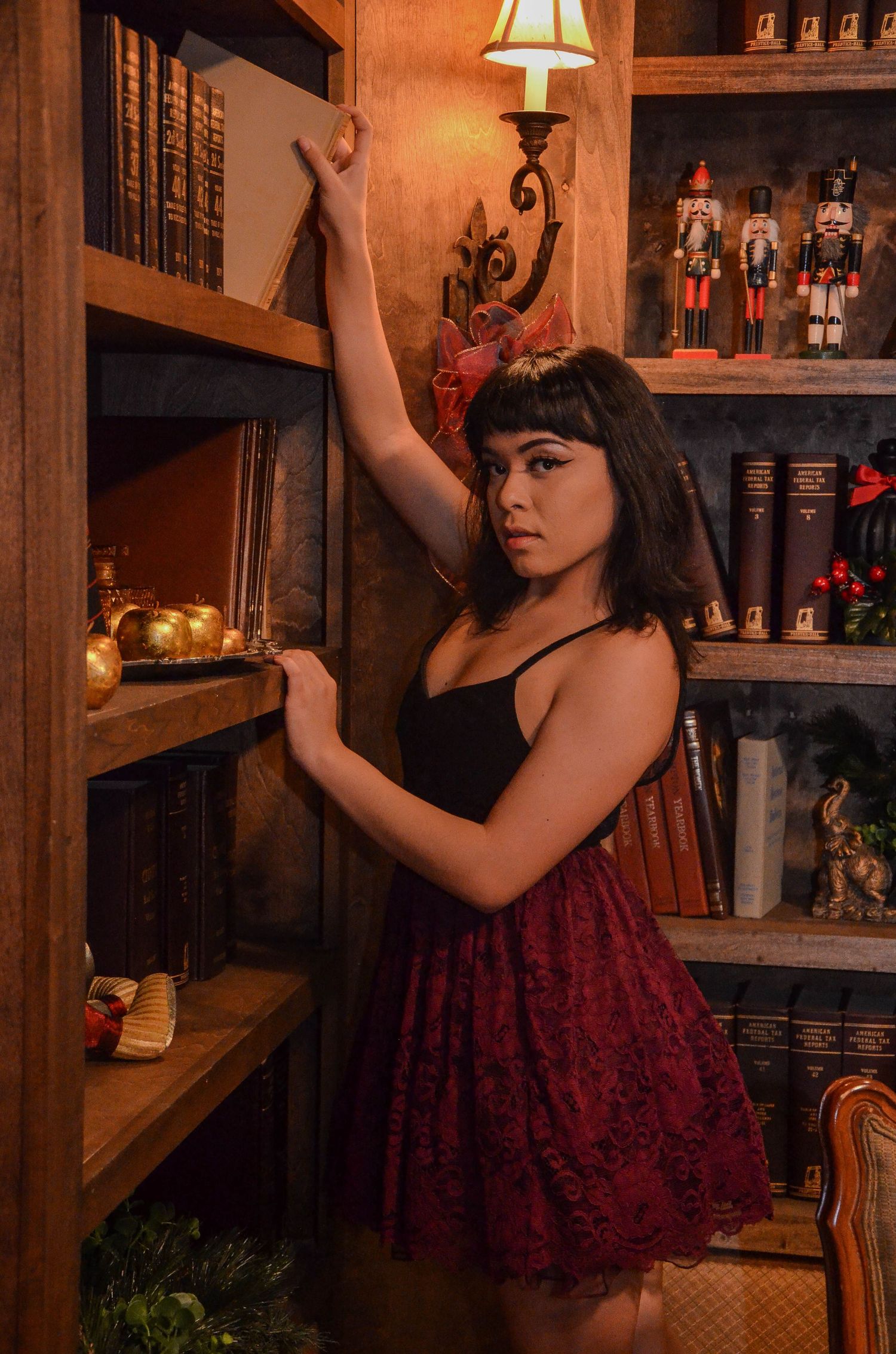 A young woman in a black top and red lace skirt posing by a wooden bookshelf with decorative items.