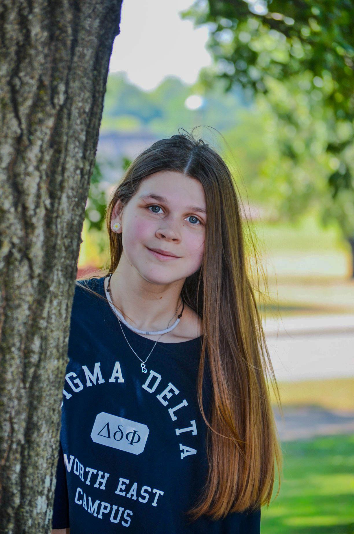 Teen girl with long brown hair leans against a tree wearing a Sigma Delta sorority t-shirt outdoors.