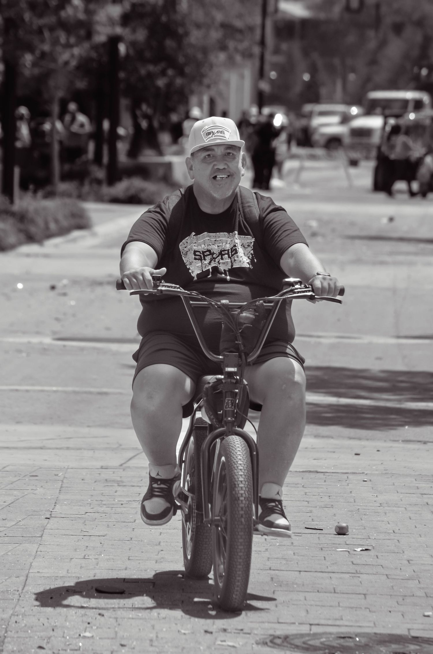 Black and white photo of overweight man riding small BMX bicycle on sunny urban sidewalk.
