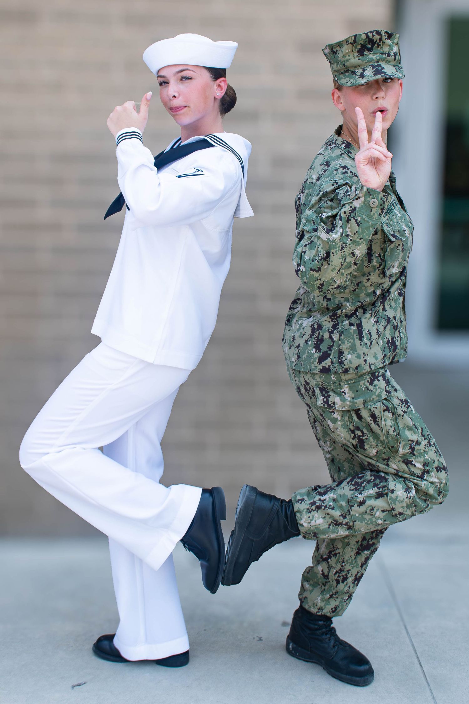 Two Navy sailors in uniform pose playfully outside a brick building.