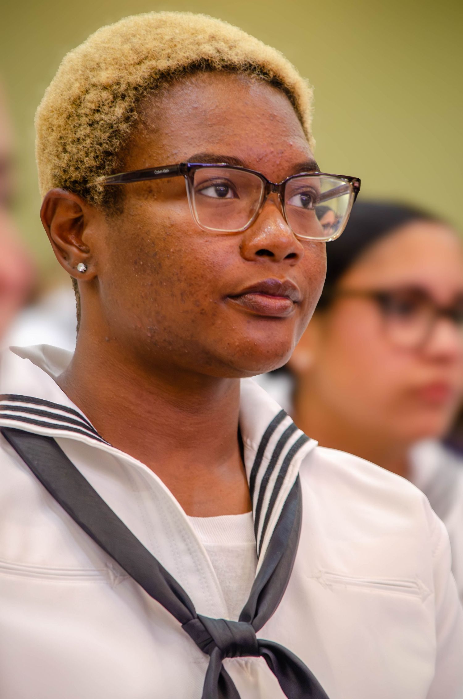 Female Navy sailor with blonde hair and glasses in white uniform at ceremony.