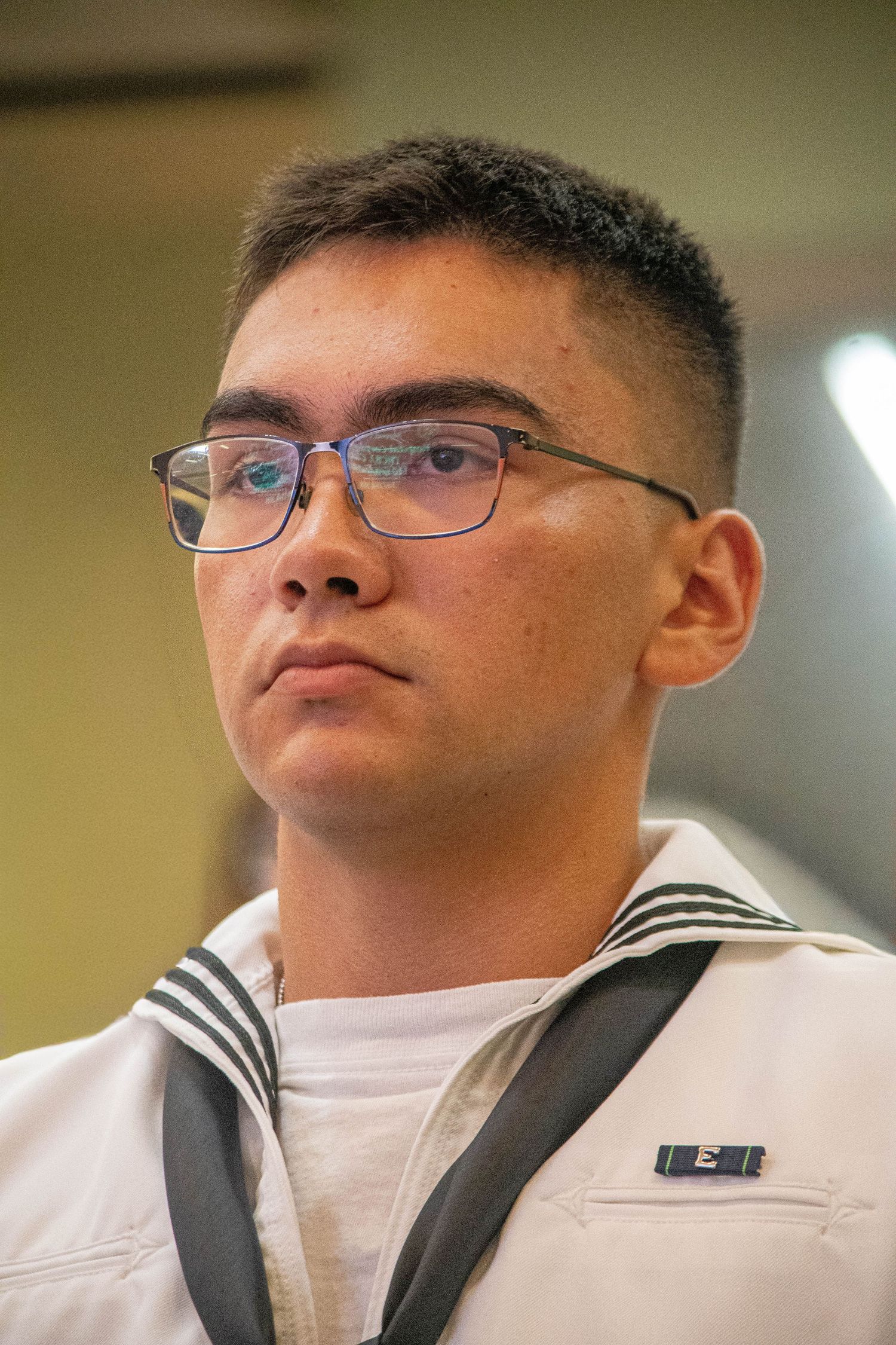 Young male Navy sailor wearing glasses in white uniform with rank insignia pin looking forward.