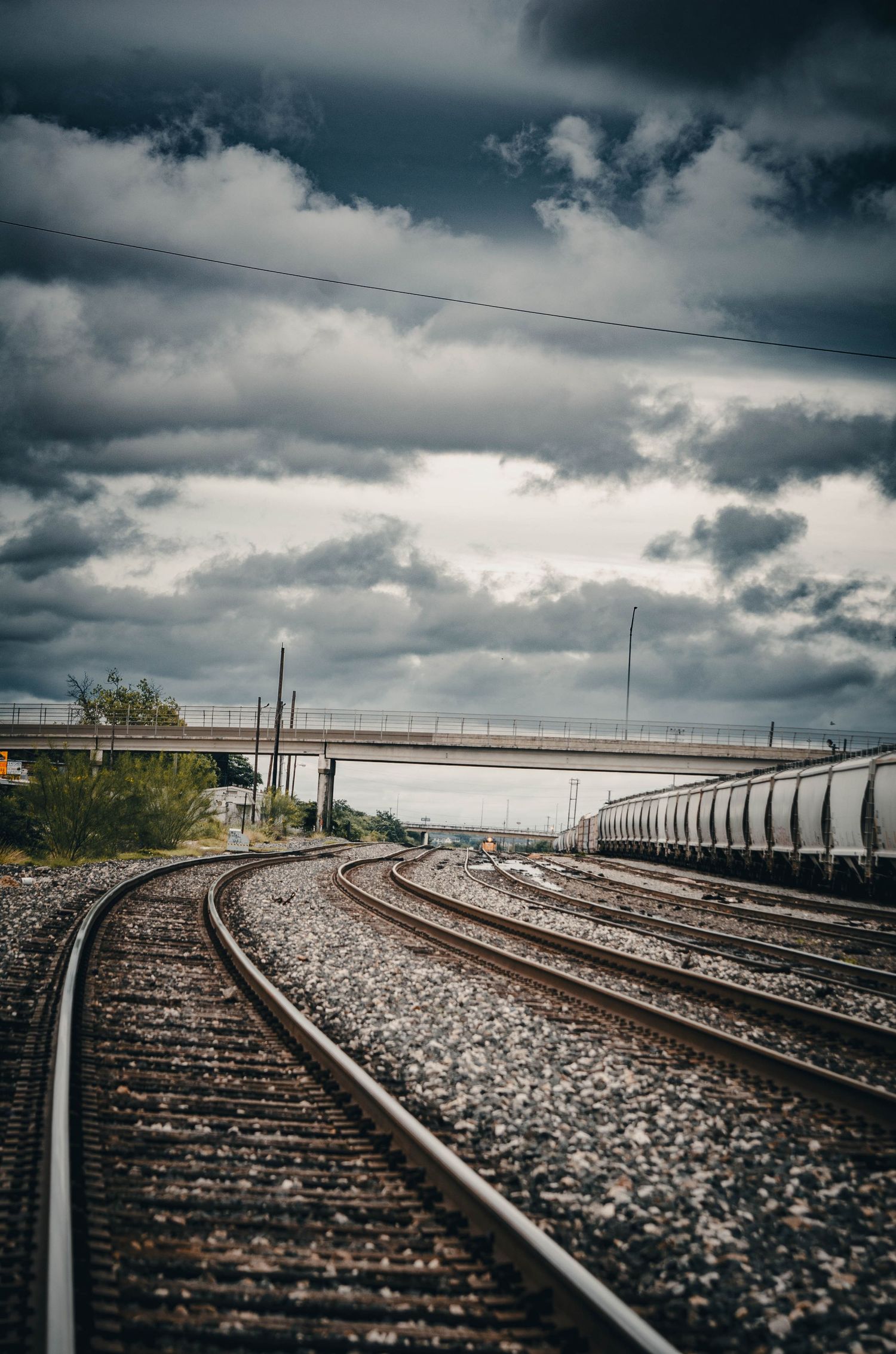 Dramatic cloudy sky over curving railroad tracks with freight train and overpass in background.