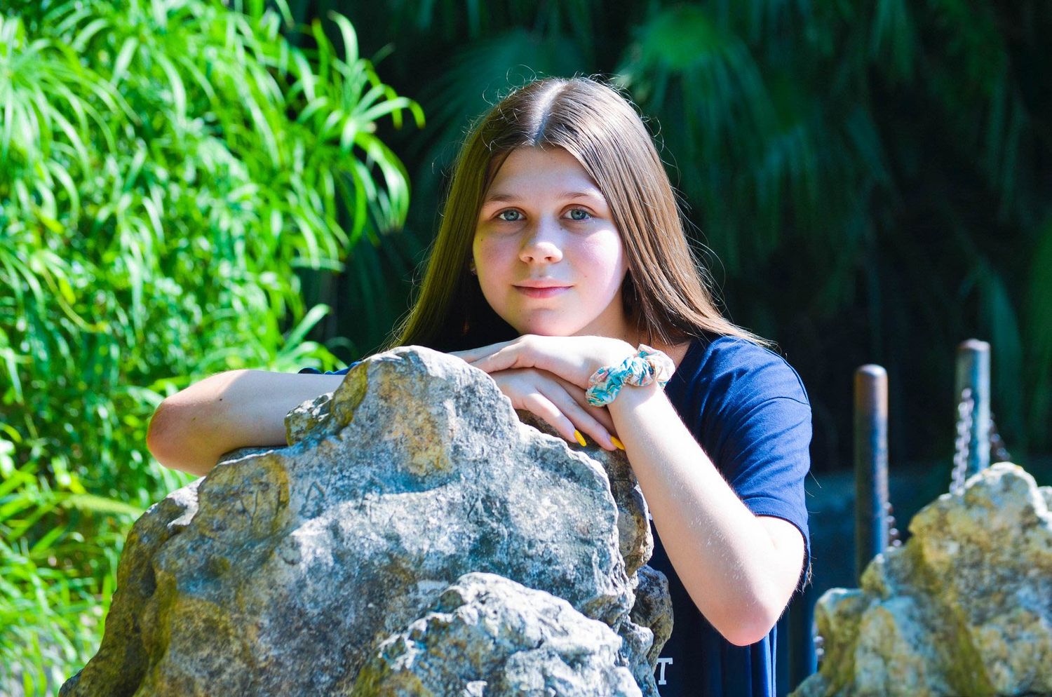 Young woman leans on a large rock in a serene garden setting with green bamboo in the background.