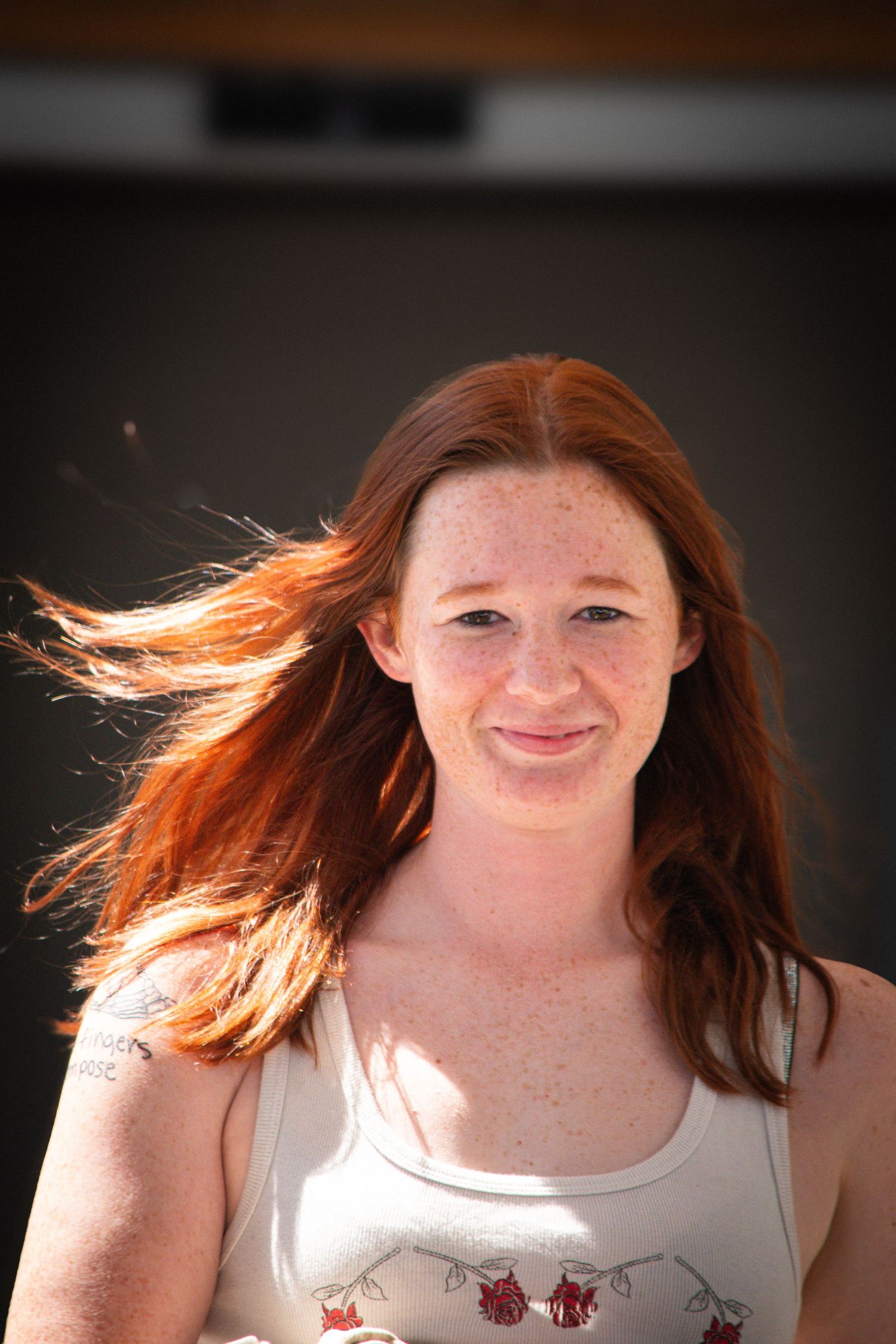 Young woman with red hair and freckles smiling outdoors, hair blowing in the wind on a sunny day.