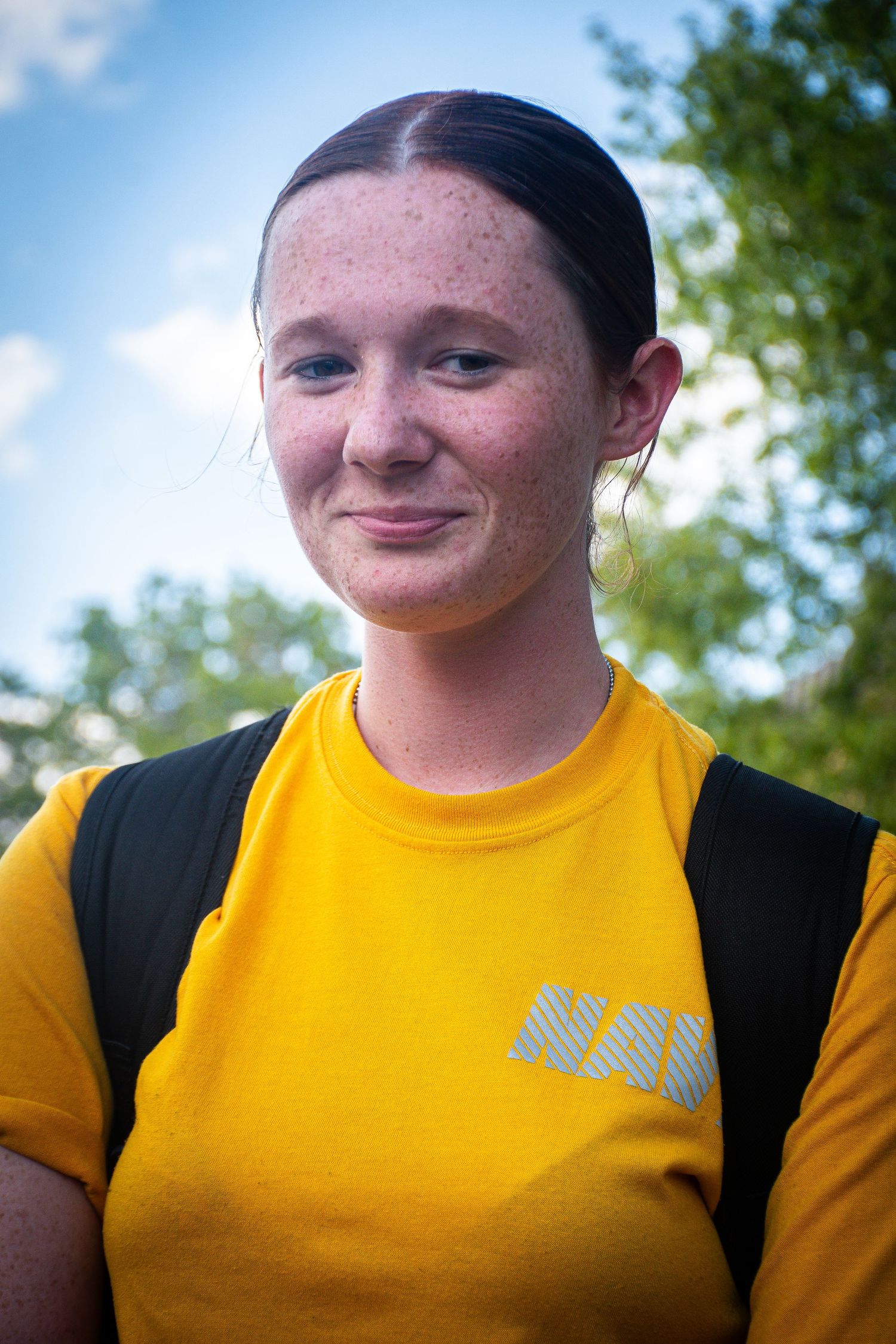 Young woman with freckles wearing a yellow NAU t-shirt and backpack, smiling outdoors with trees behind her.