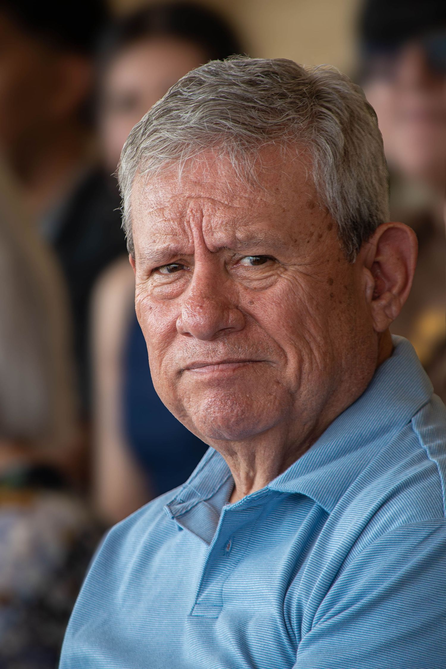 An older man with gray hair in a light blue polo shirt looks at the camera seriously.