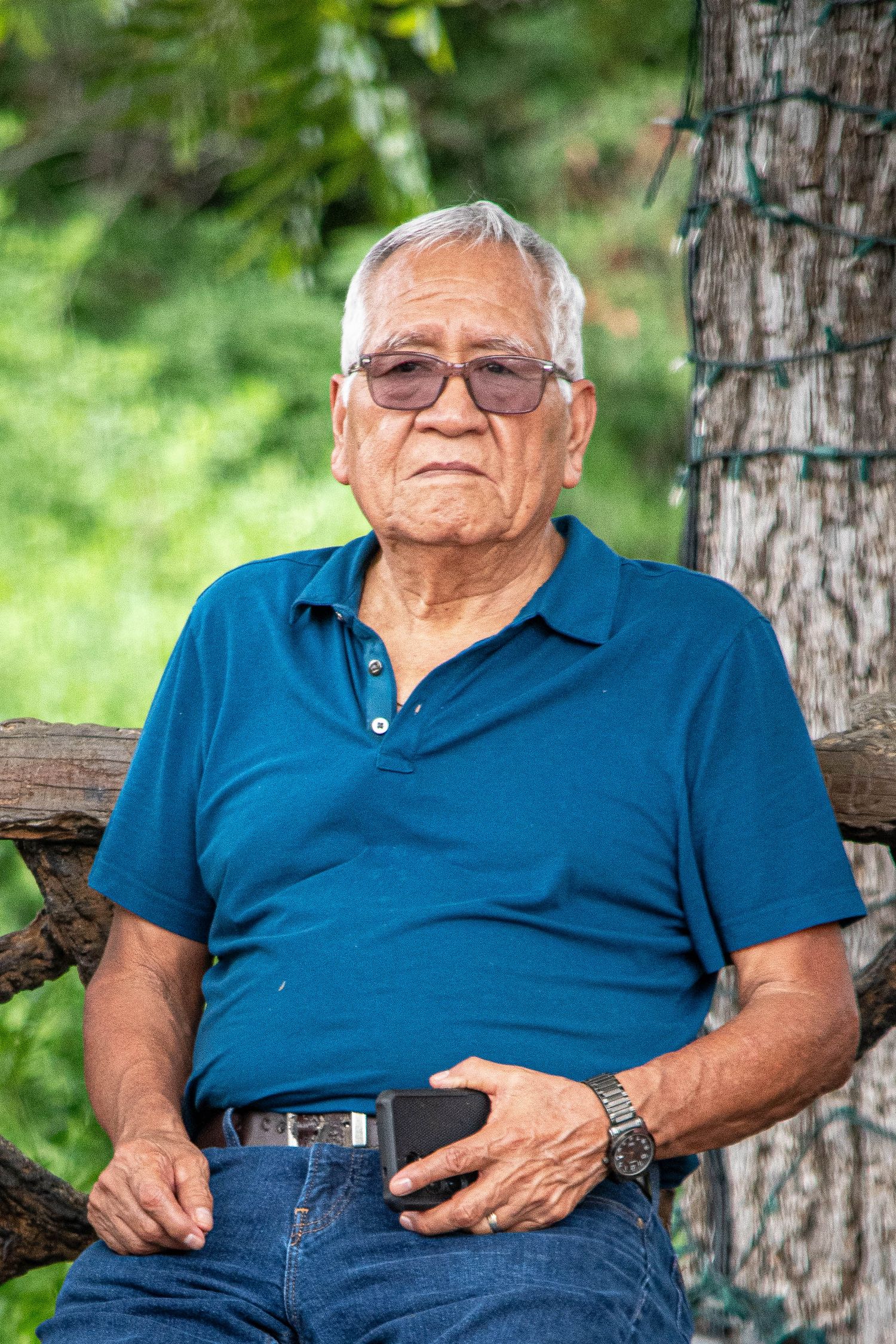 Elderly man with white hair and glasses in blue polo shirt sitting on wooden bench.
