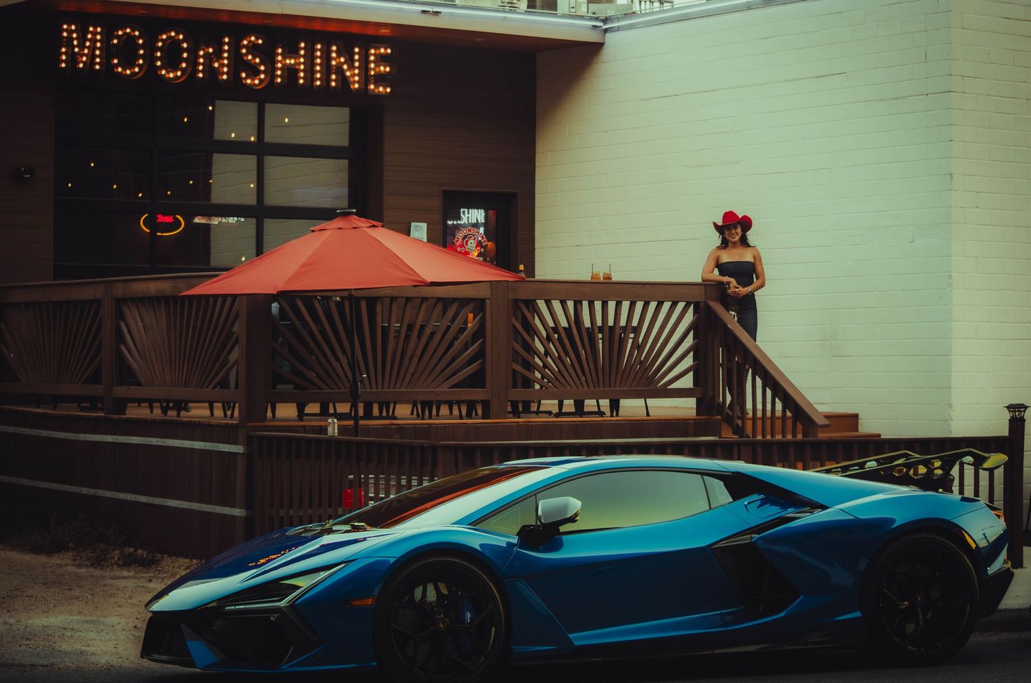 Blue Lamborghini supercar parked outside the Moonshine restaurant with a woman standing on deck.