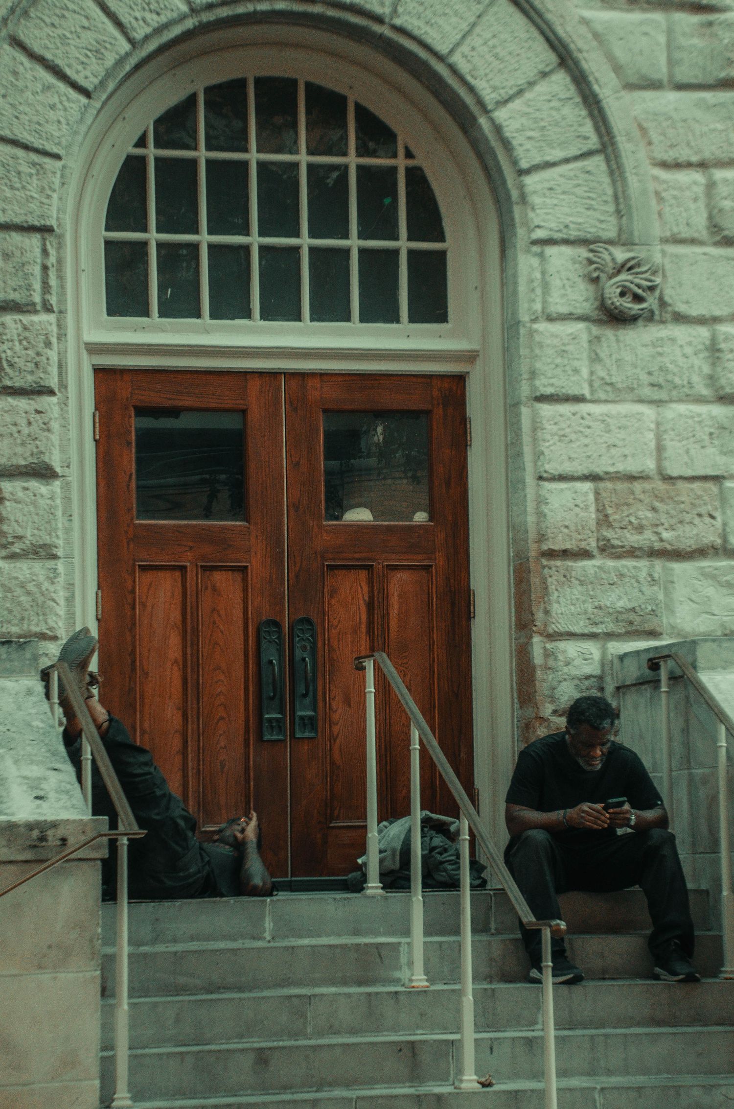 Two people sit on steps outside a historic stone building with arched wooden double doors.