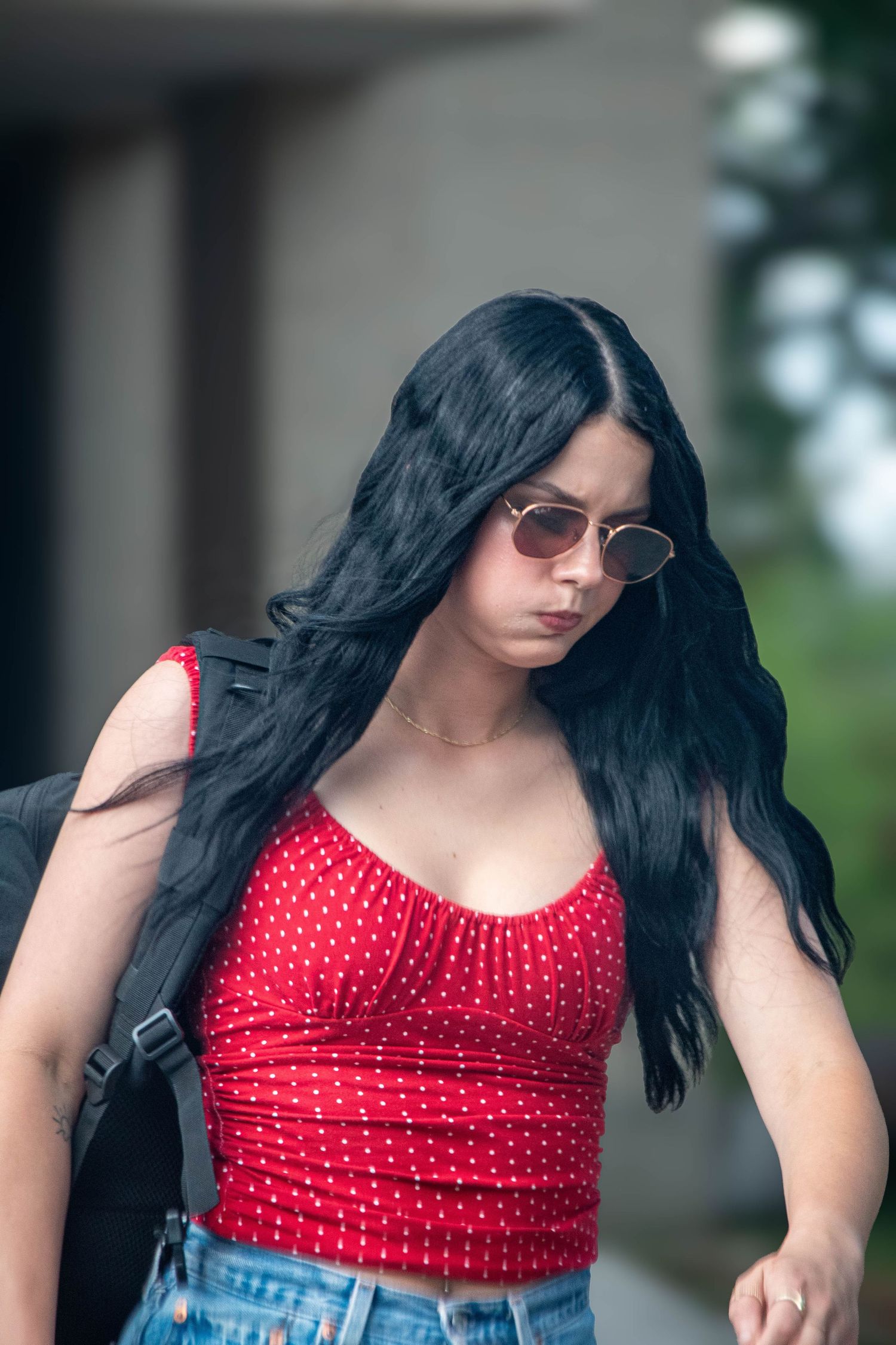 Young woman with long black hair wearing red polka dot crop top, sunglasses, and carrying a backpack outdoors.