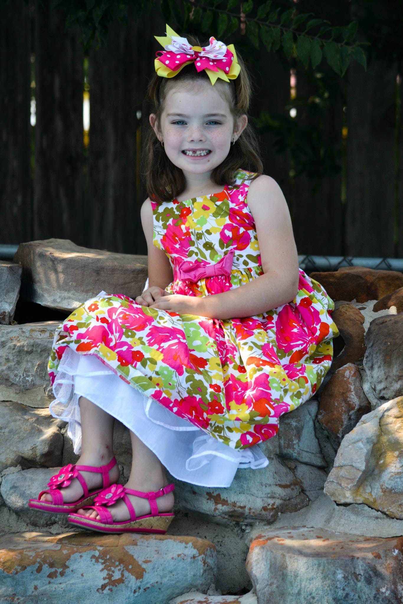 Happy young girl in colorful floral dress and pink sandals sitting on rocks.