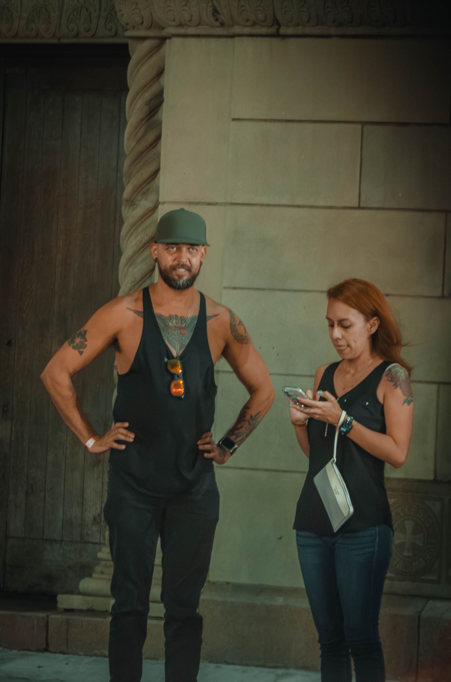 A tattooed muscular man in a tank top and cap stands beside a woman using her smartphone outdoors.
