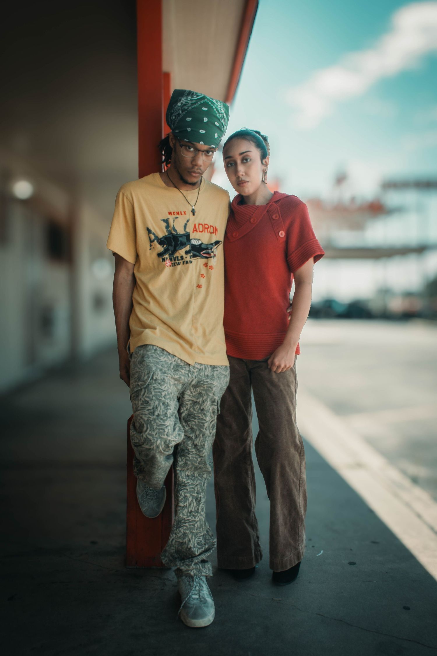 Two young people in retro streetwear pose together outside a motel, wearing earth tones and patterned pants.