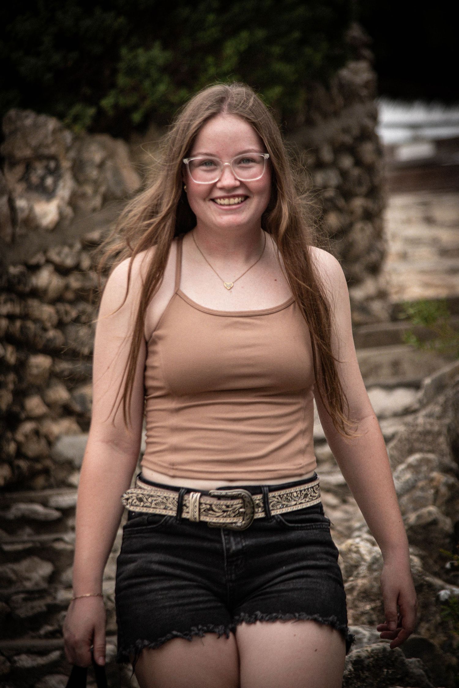 Smiling young woman with glasses and long hair wearing a tan tank top and black denim shorts outdoors.