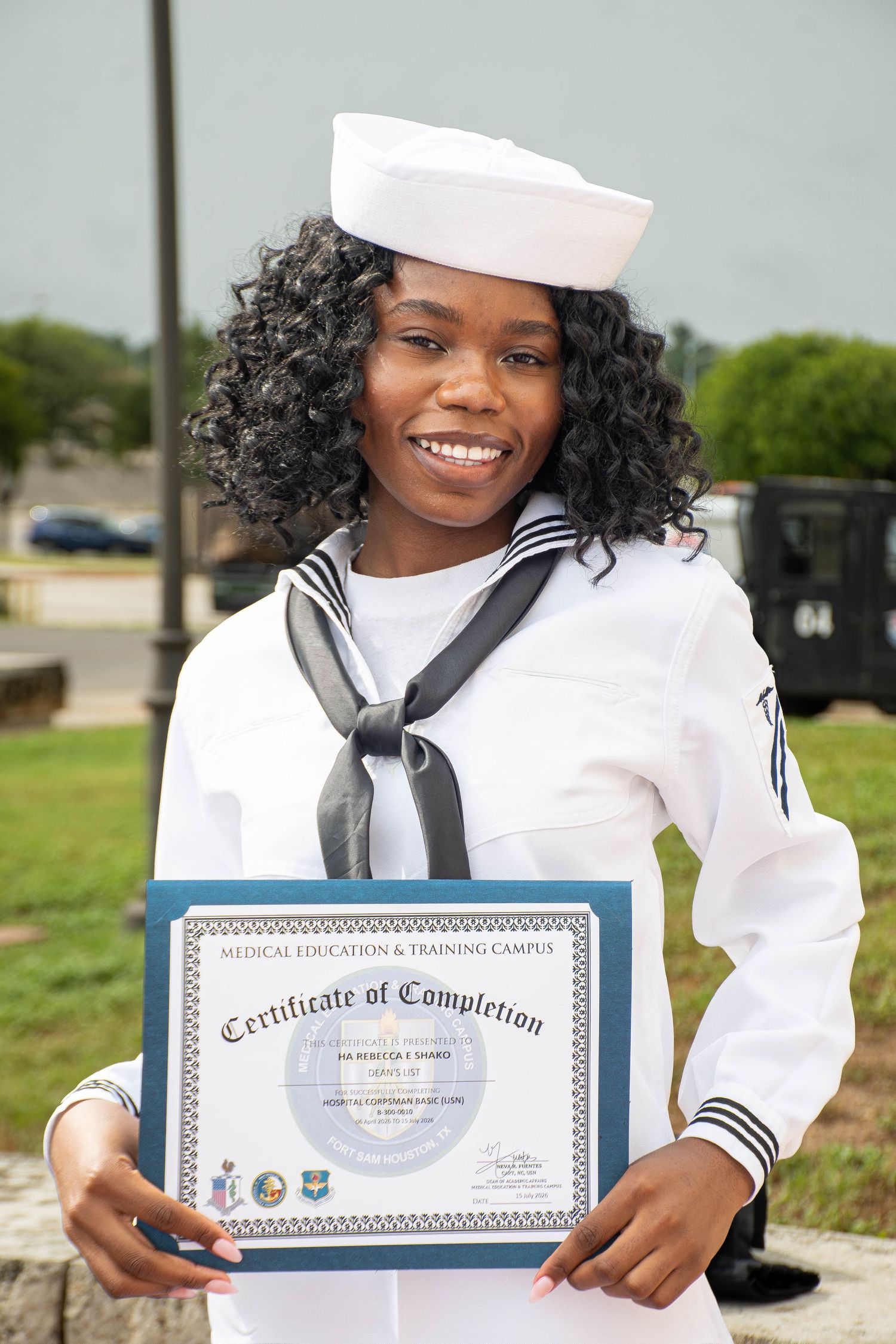 Smiling female Navy sailor in dress whites proudly holds Certificate of Completion outdoors.