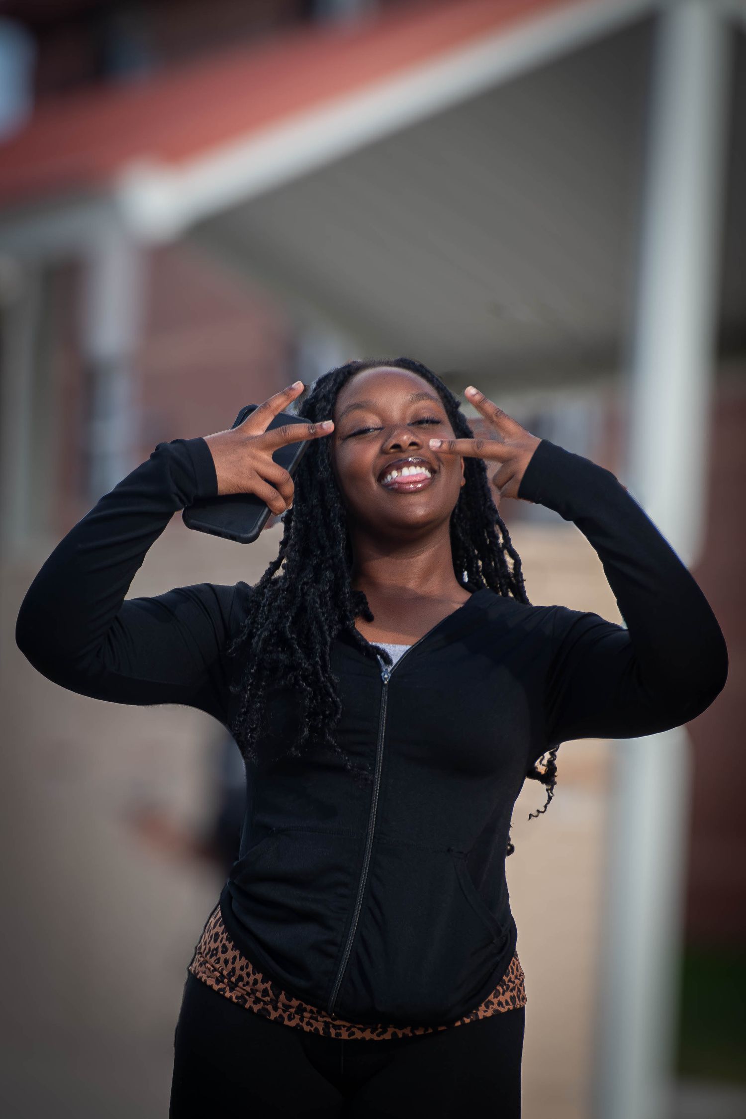 Young Black woman with braids in black zip-up jacket makes peace signs near face, smiling outdoors near brick building.